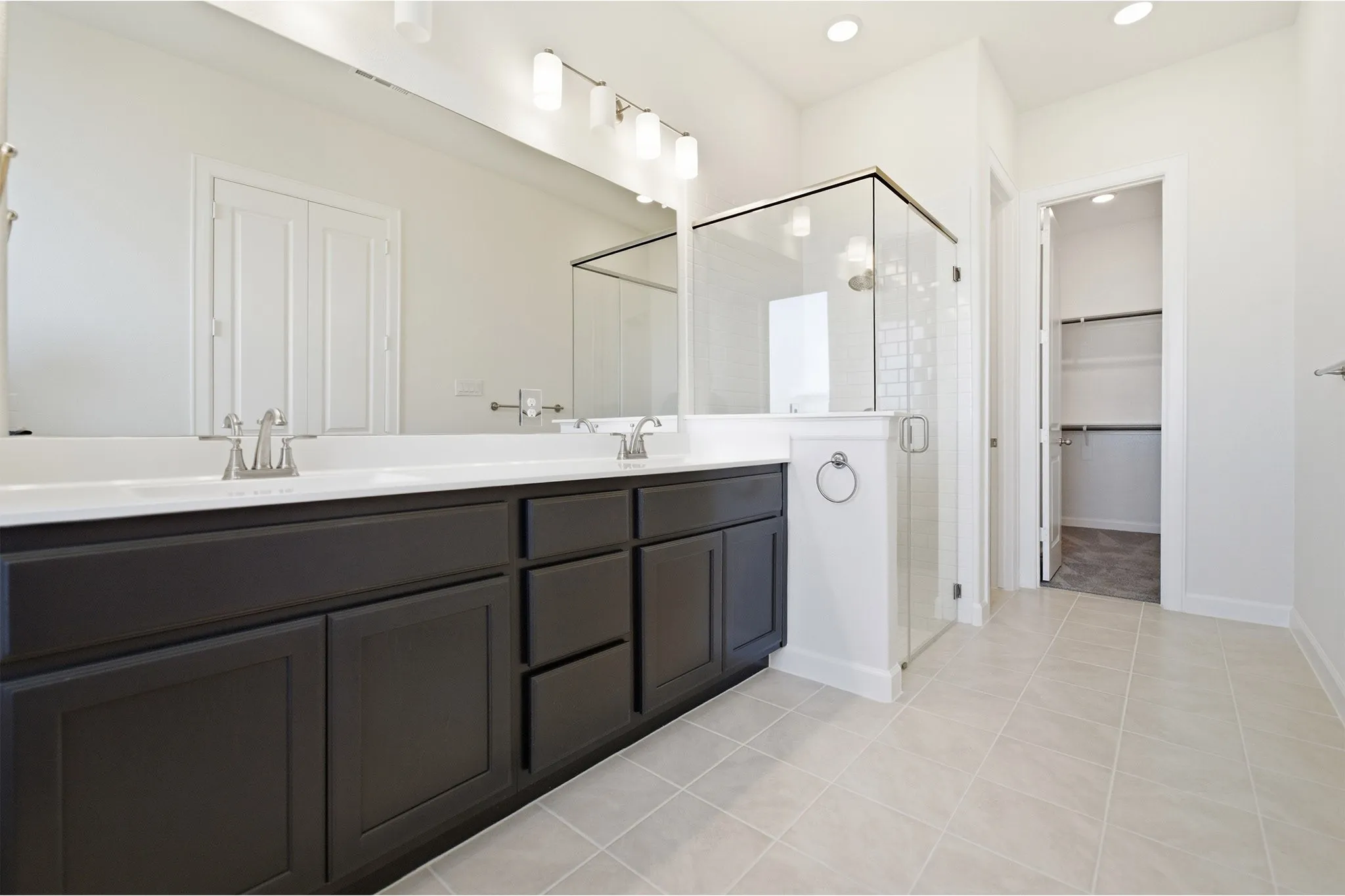 Bathroom featuring a dual vanity with dark wood-finish cabinetry, a large frameless mirror, a glass-enclosed shower with subway tile, and light-toned floor tiling