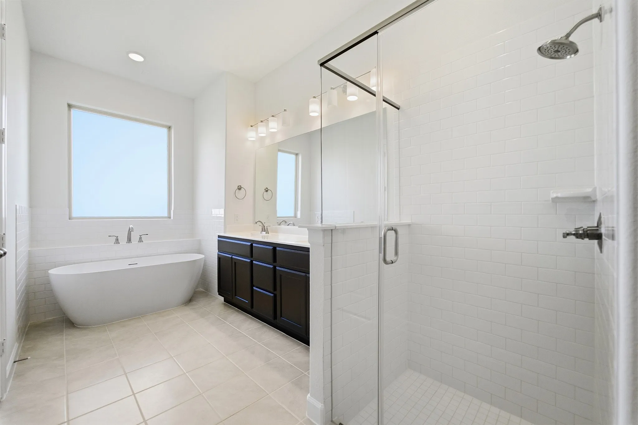 Spa-like bathroom featuring a freestanding tub, glass-enclosed shower with subway tile, a double vanity with dark cabinetry, and light tile flooring