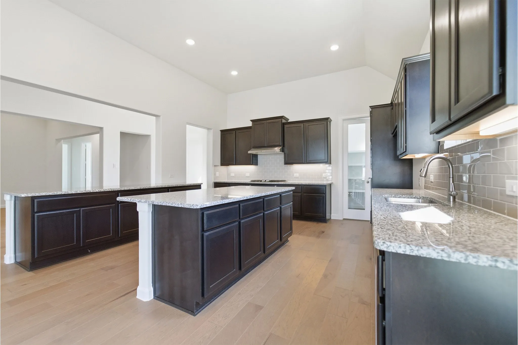 Open-concept kitchen featuring dark wood cabinetry, light granite countertops, a subway tile backsplash, a stainless steel sink with a gooseneck faucet, and light wood-finish flooring