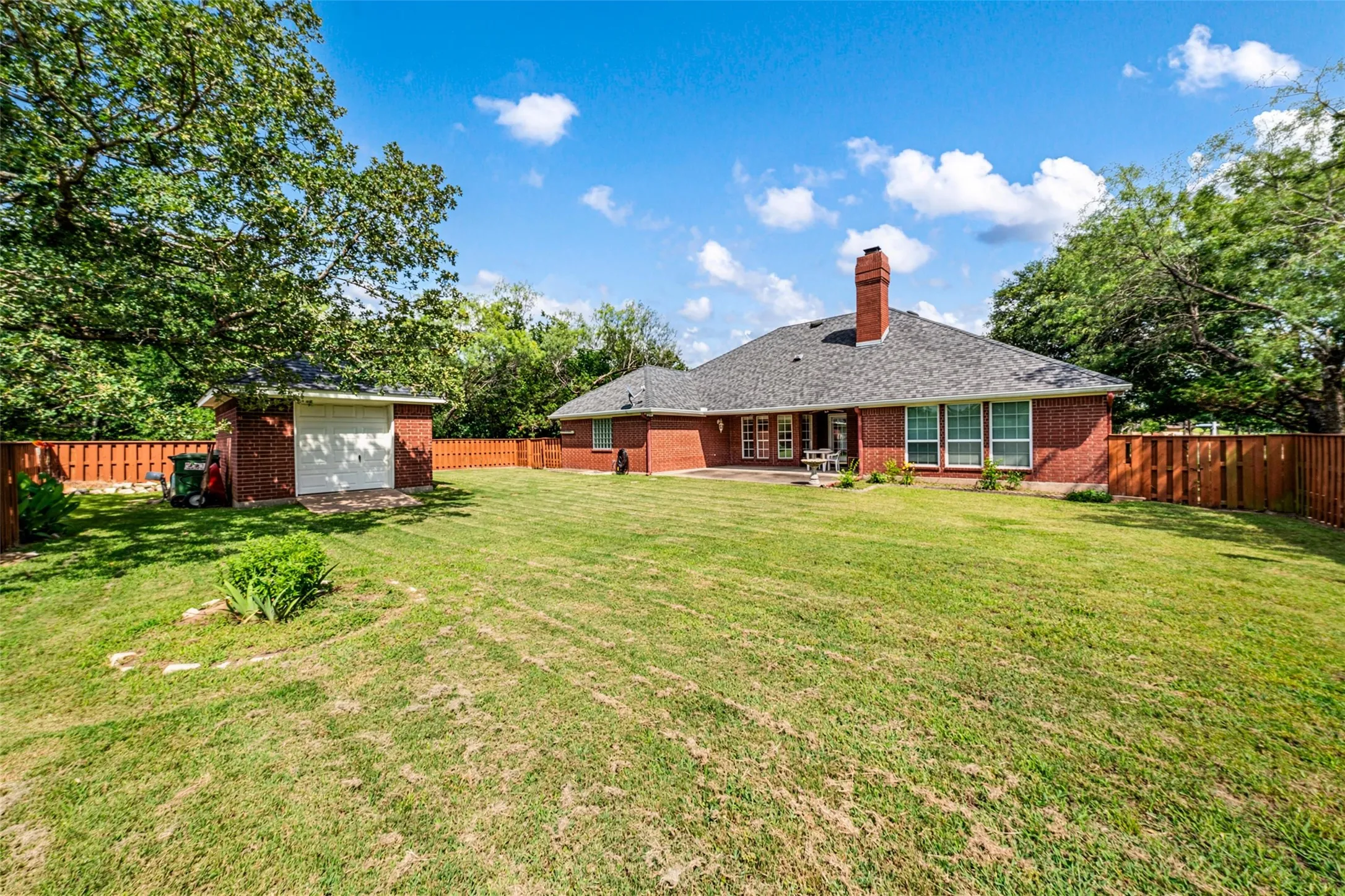 Expansive backyard featuring a manicured lawn, a detached brick storage building with a garage door, a wooden privacy fence, and mature trees
