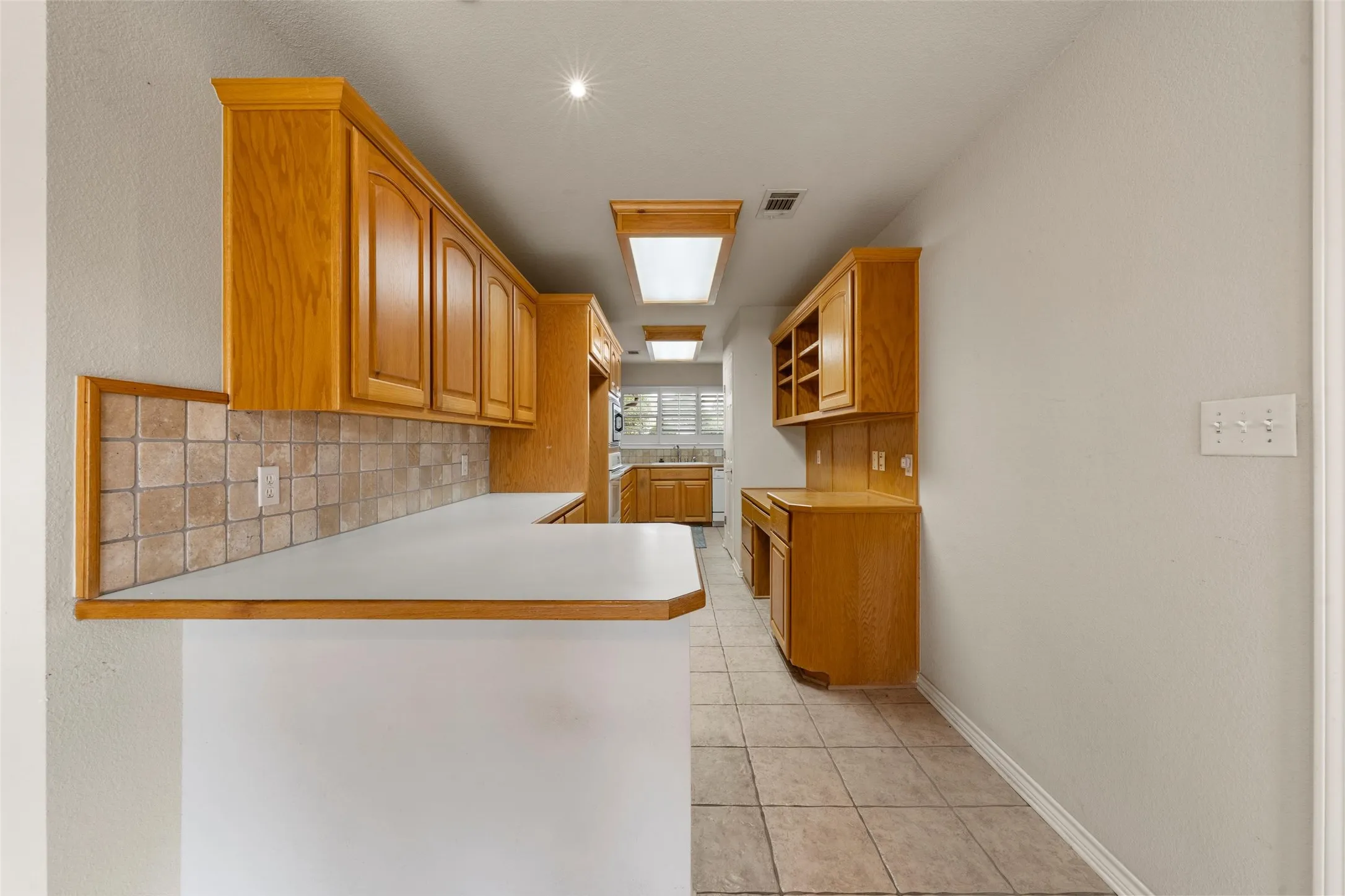 Galley kitchen featuring wood-finish cabinetry, tile flooring, light countertops, and a tiled backsplash