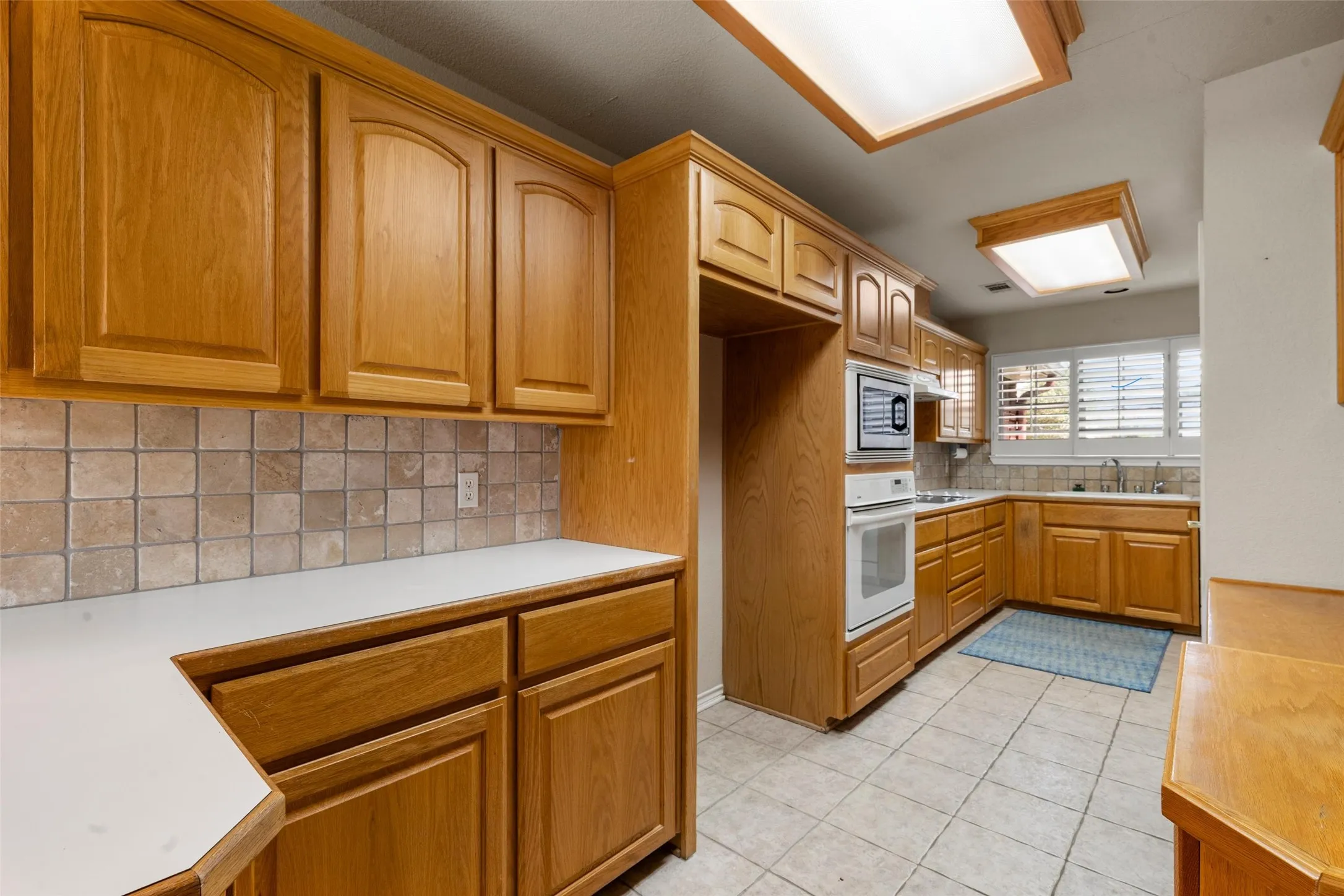Kitchen featuring wood cabinetry, solid surface countertops, tile backsplash, tile flooring, and integrated white appliances