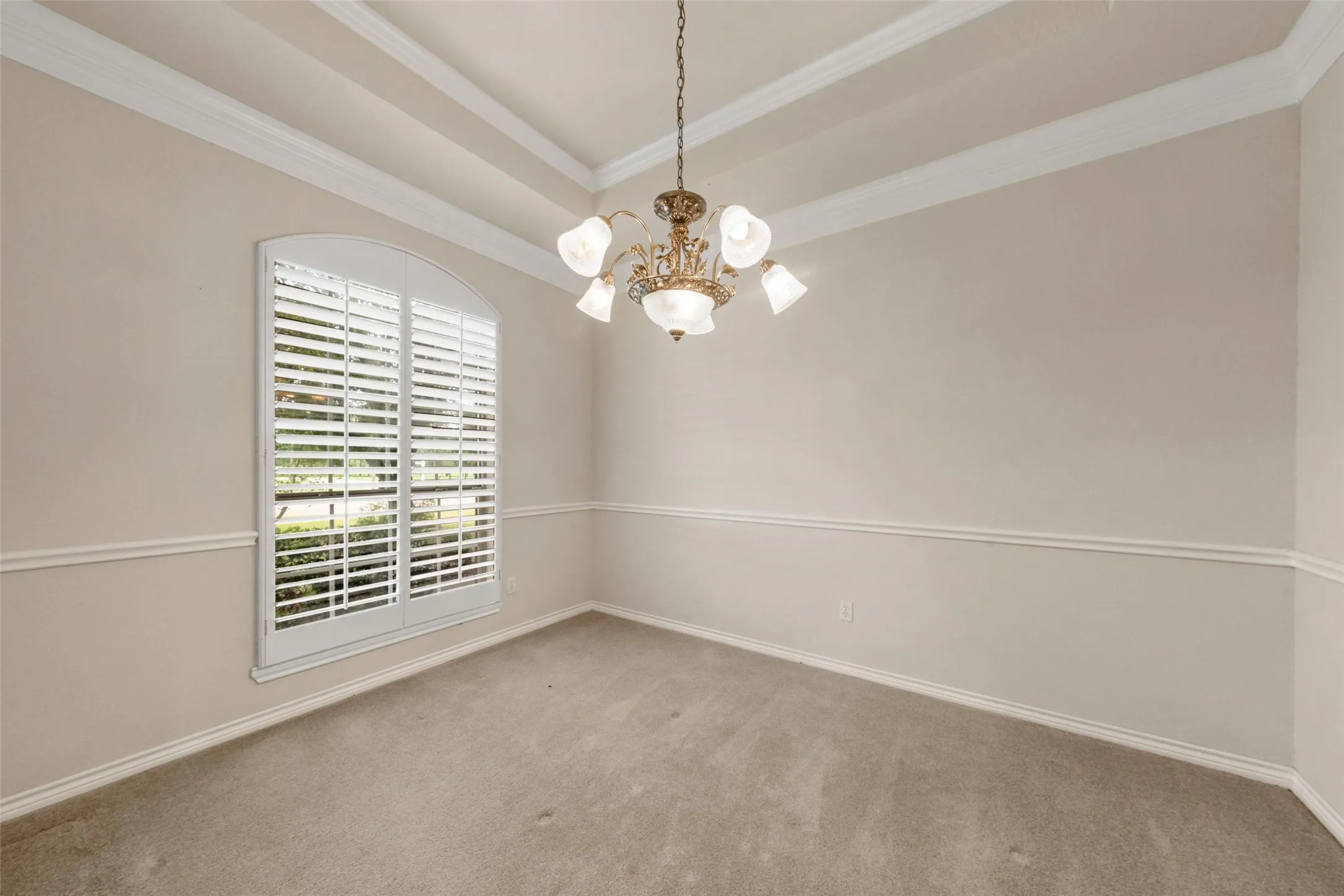 Arched window with plantation shutters, featuring a coffered ceiling, elaborate chandelier, chair rail molding, and neutral carpeting