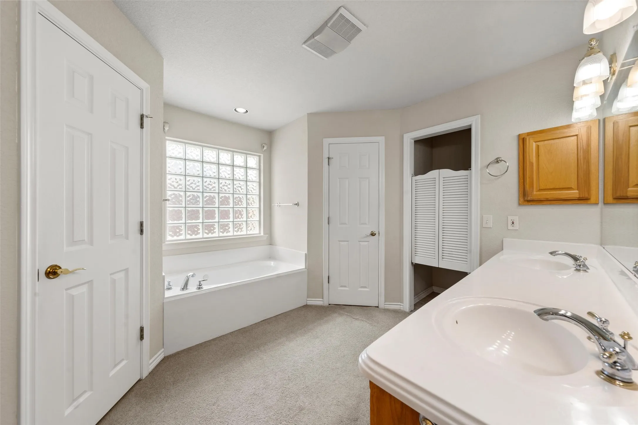 Double vanity with white countertops and chrome fixtures, built-in bathtub with glass block window, and light wood cabinetry