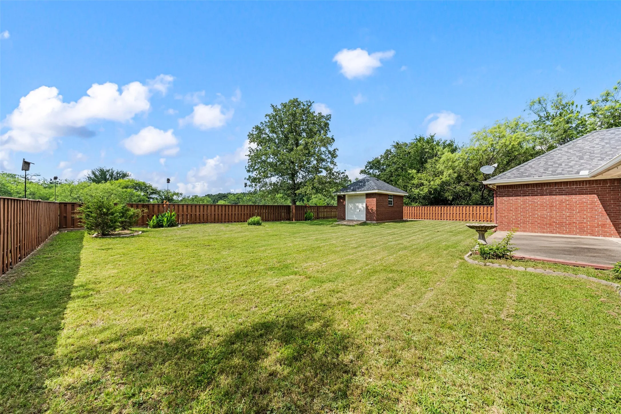Expansive rear yard featuring a privacy fence, mature trees, a brick storage shed, and a concrete patio