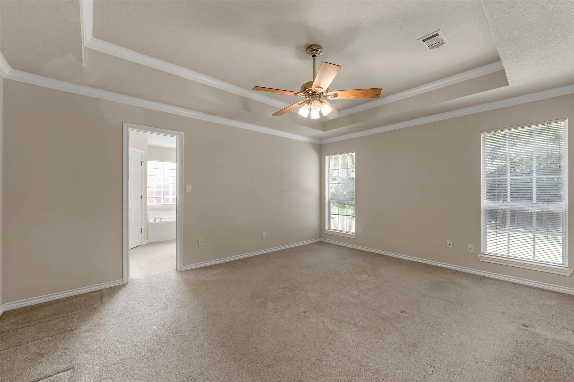 Spacious bedroom featuring a tray ceiling with crown molding, two large windows, a ceiling fan, and carpet flooring