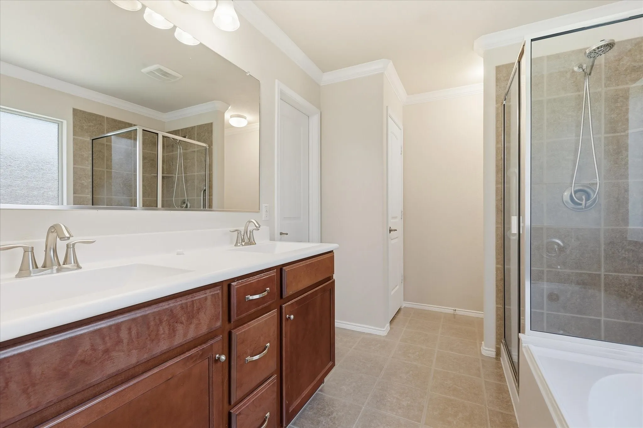 Bathroom featuring a double vanity with wood-finish cabinetry and brushed nickel hardware, a large vanity mirror, a frosted window, a glass-enclosed shower with tiled walls, and light-toned tile flooring