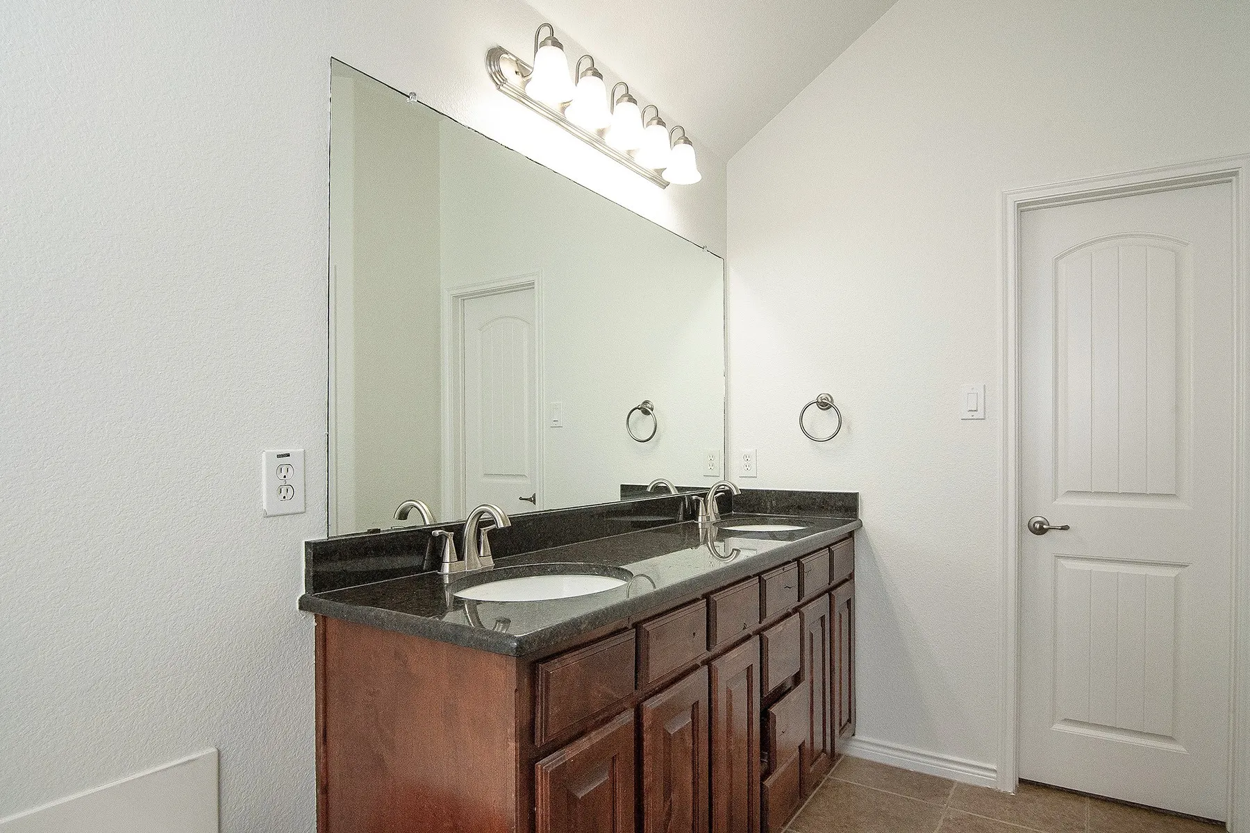 Master bathroom vanity featuring rich wood cabinetry with dual sinks, dark countertop surface, polished chrome fixtures, and an illuminated mirror with sconce lighting above