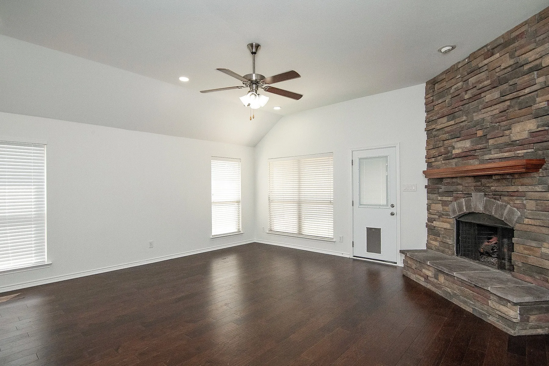 Spacious living area featuring rich wood-finish flooring, ceiling fan with integrated lighting, and multiple windows providing natural light