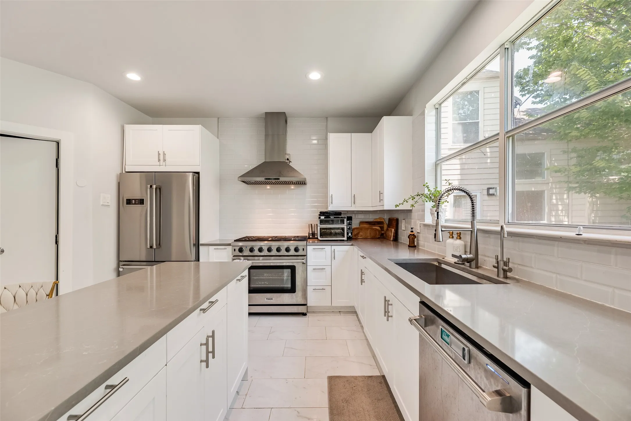 Modern kitchen featuring an extended island with ample storage, stainless steel appliances, a range hood, and a large window above the sink
