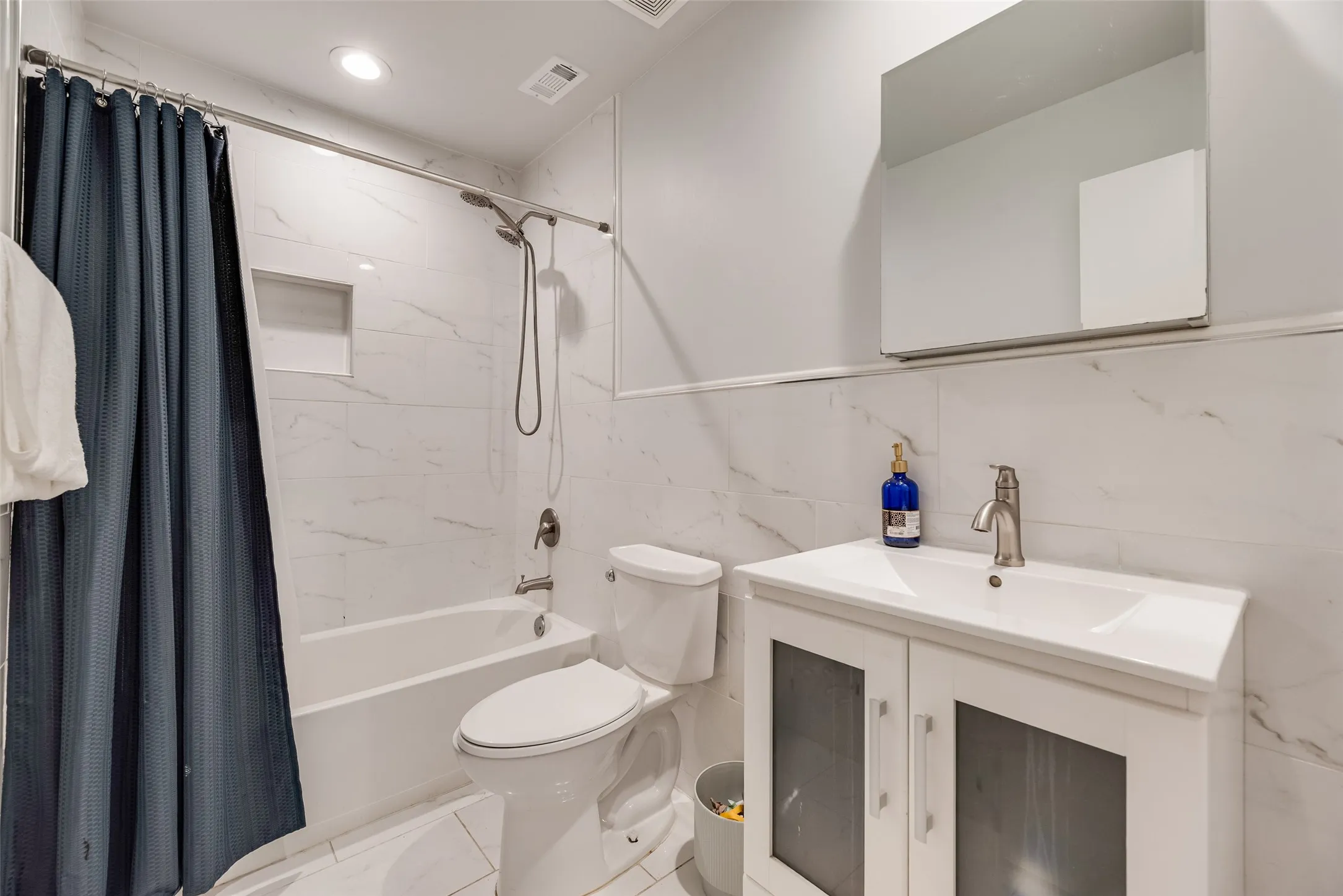 Bathroom featuring full white subway tile surrounding the tub, a built-in shower niche, a white vanity with frosted glass doors, and a rectangular mirror