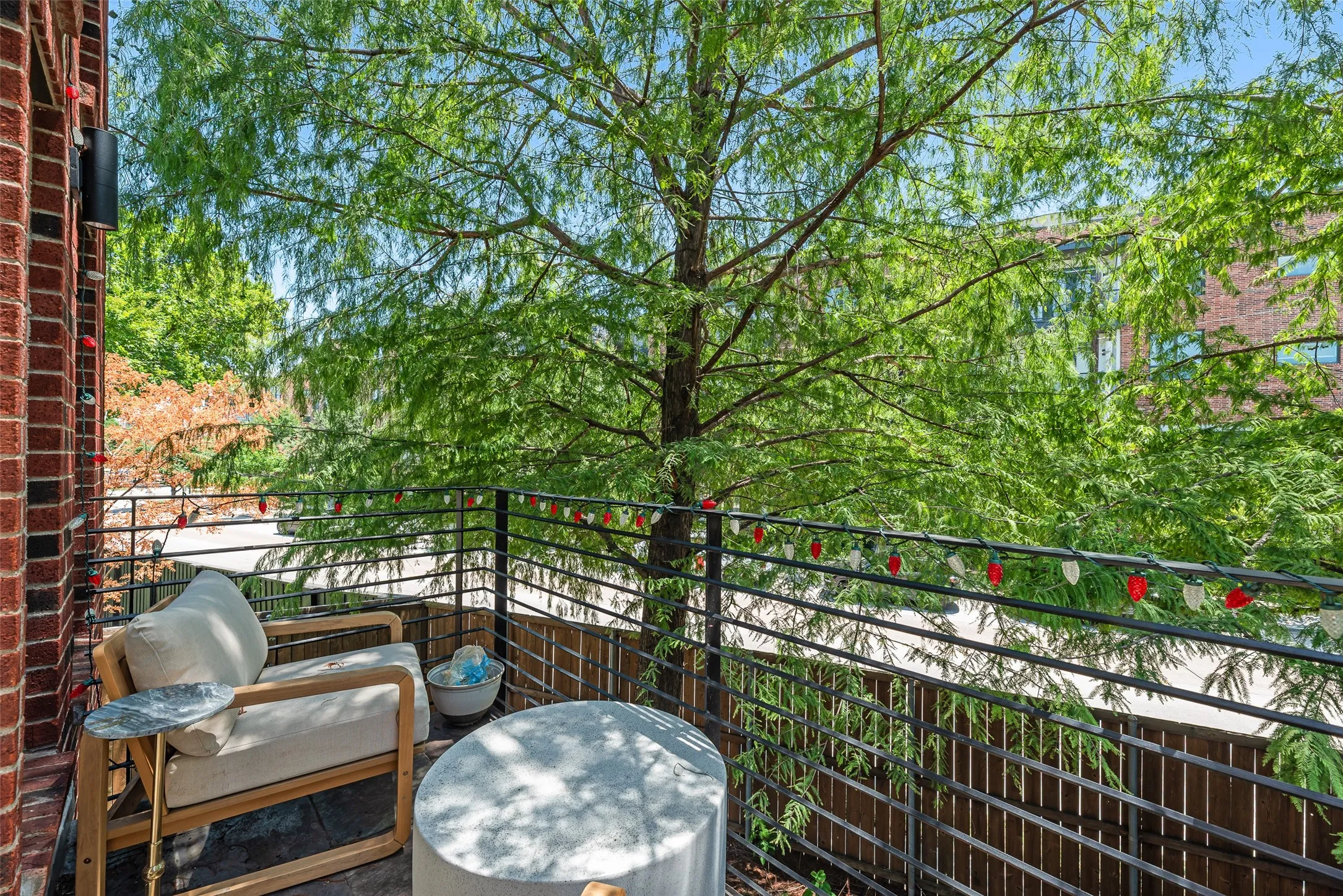 Exterior balcony featuring brick siding, a black metal railing, and a mature tree providing natural shade