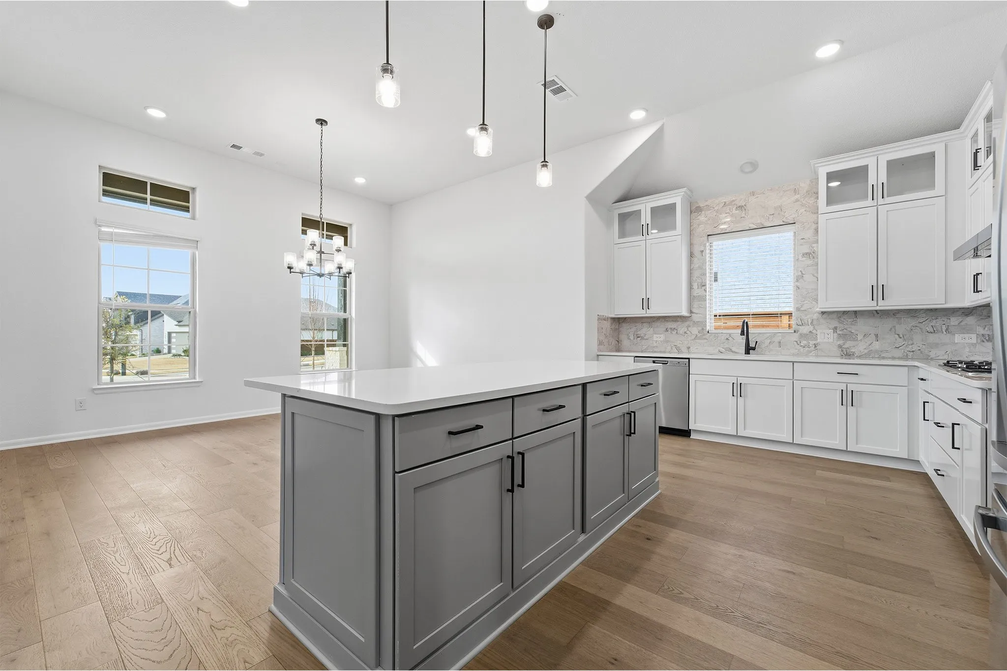 Kitchen featuring a large central island with a light-toned countertop, wood-finish flooring, white cabinetry with black hardware, a light-toned tile backsplash, and stainless steel​​‌​​​​‌​​‌‌​‌‌​​​‌‌​‌​‌​‌​​​‌​​ appliances