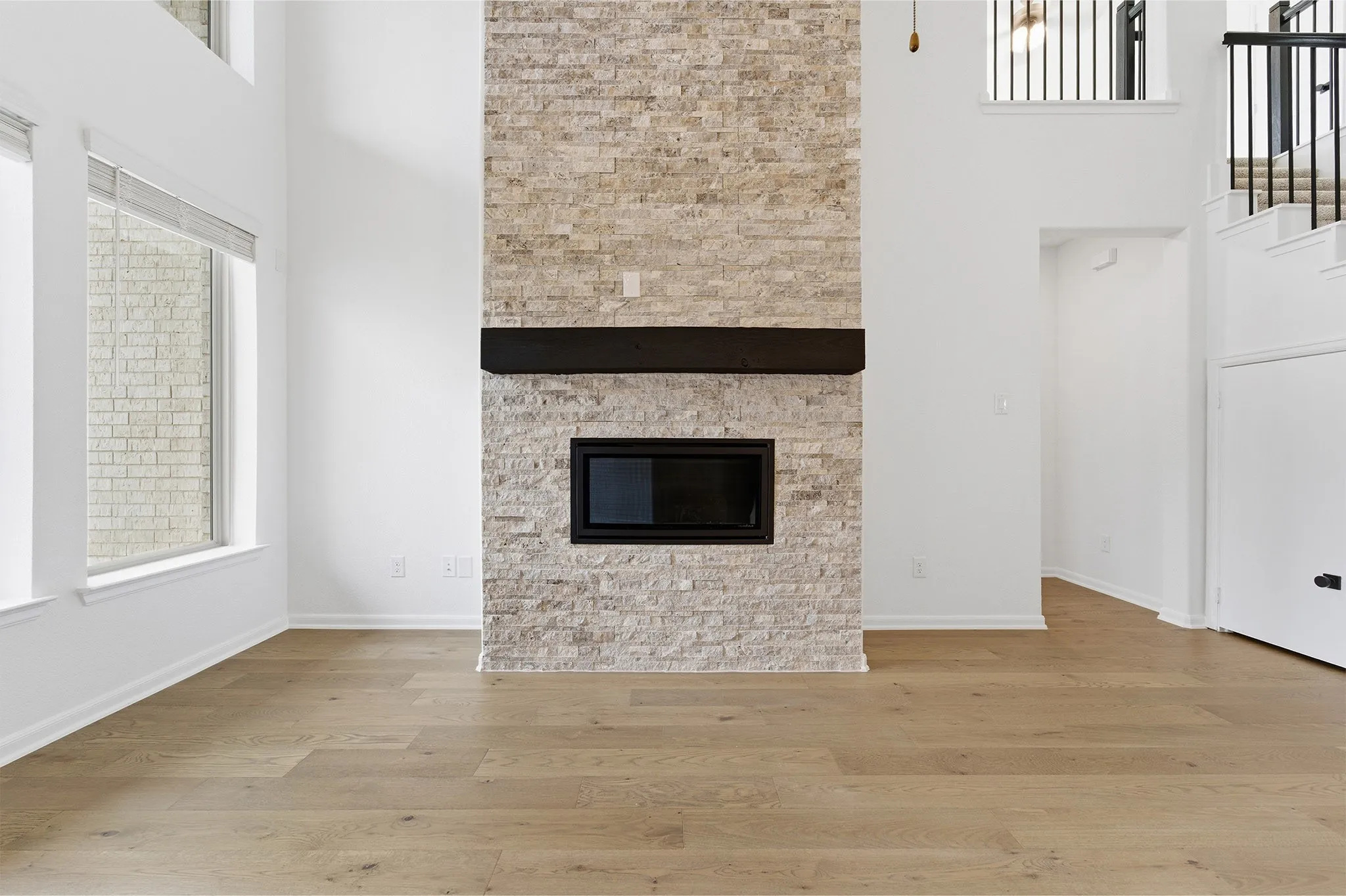 Two-story living area featuring a stacked stone fireplace with a dark wood mantle, wood-finish flooring, and multiple large windows with blinds
