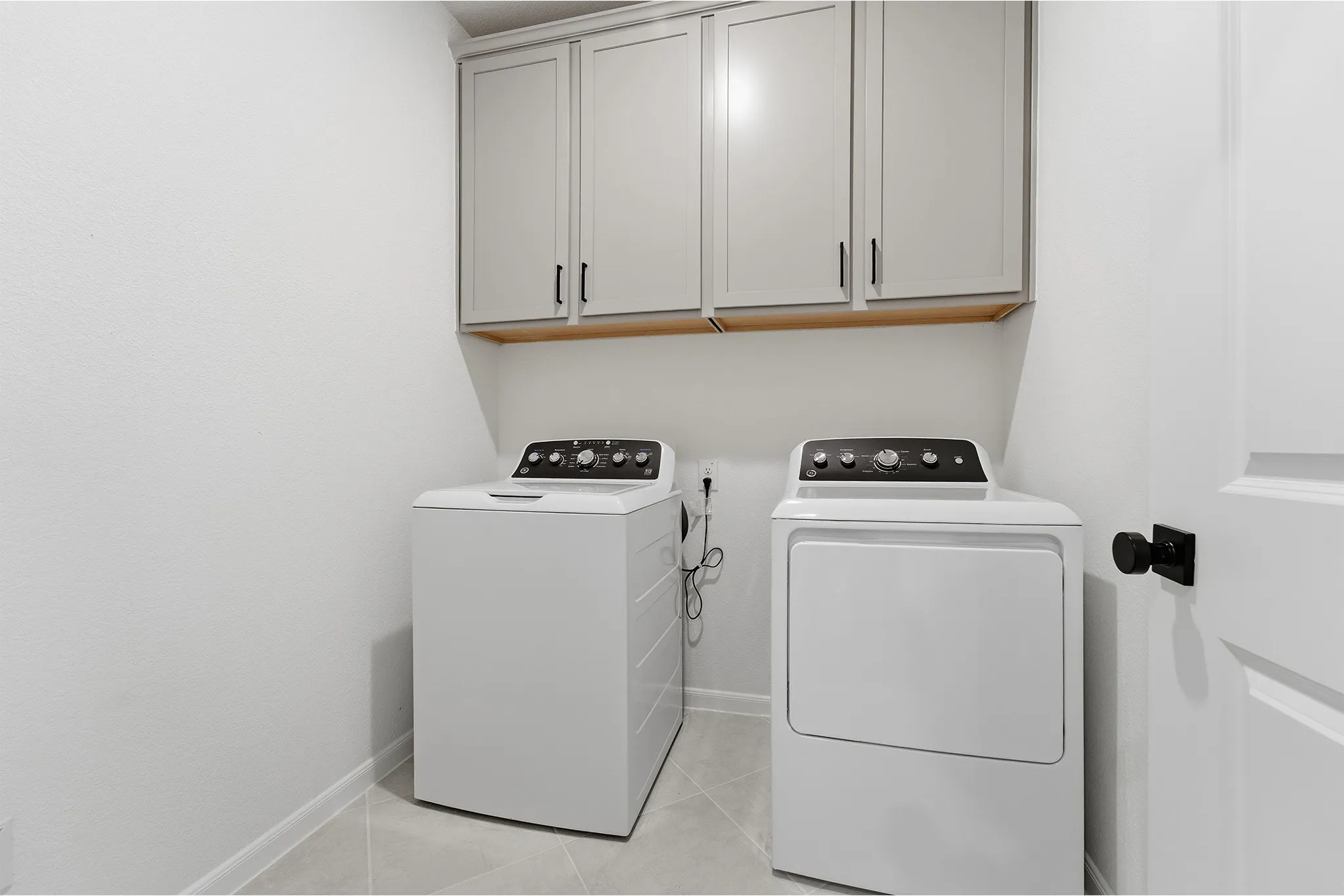 Dedicated laundry area featuring light gray cabinetry with black hardware, light wood-finish shelving, and light gray tiled flooring