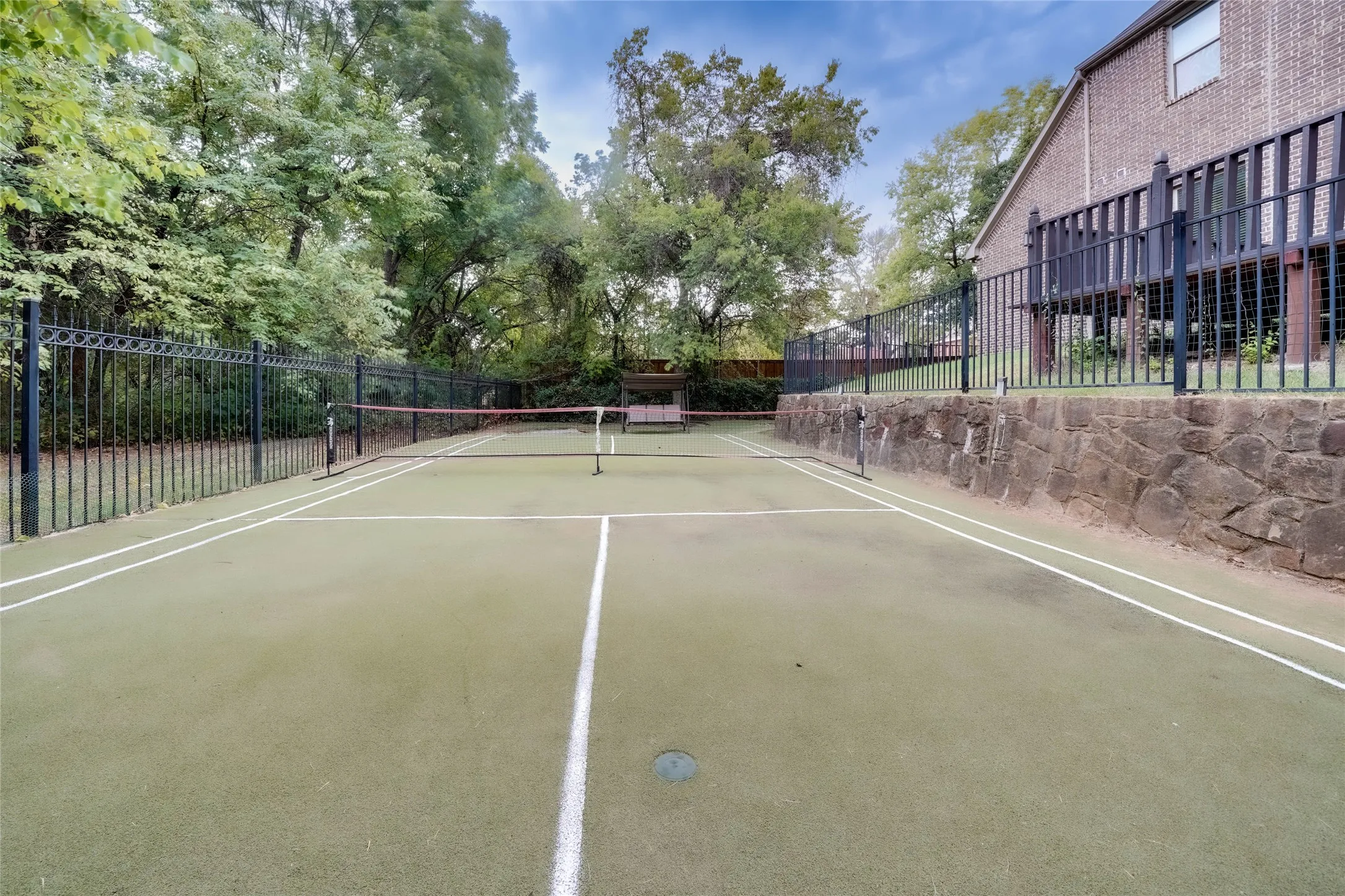 Private tennis court featuring a green surface with white lines, surrounded by a black metal fence