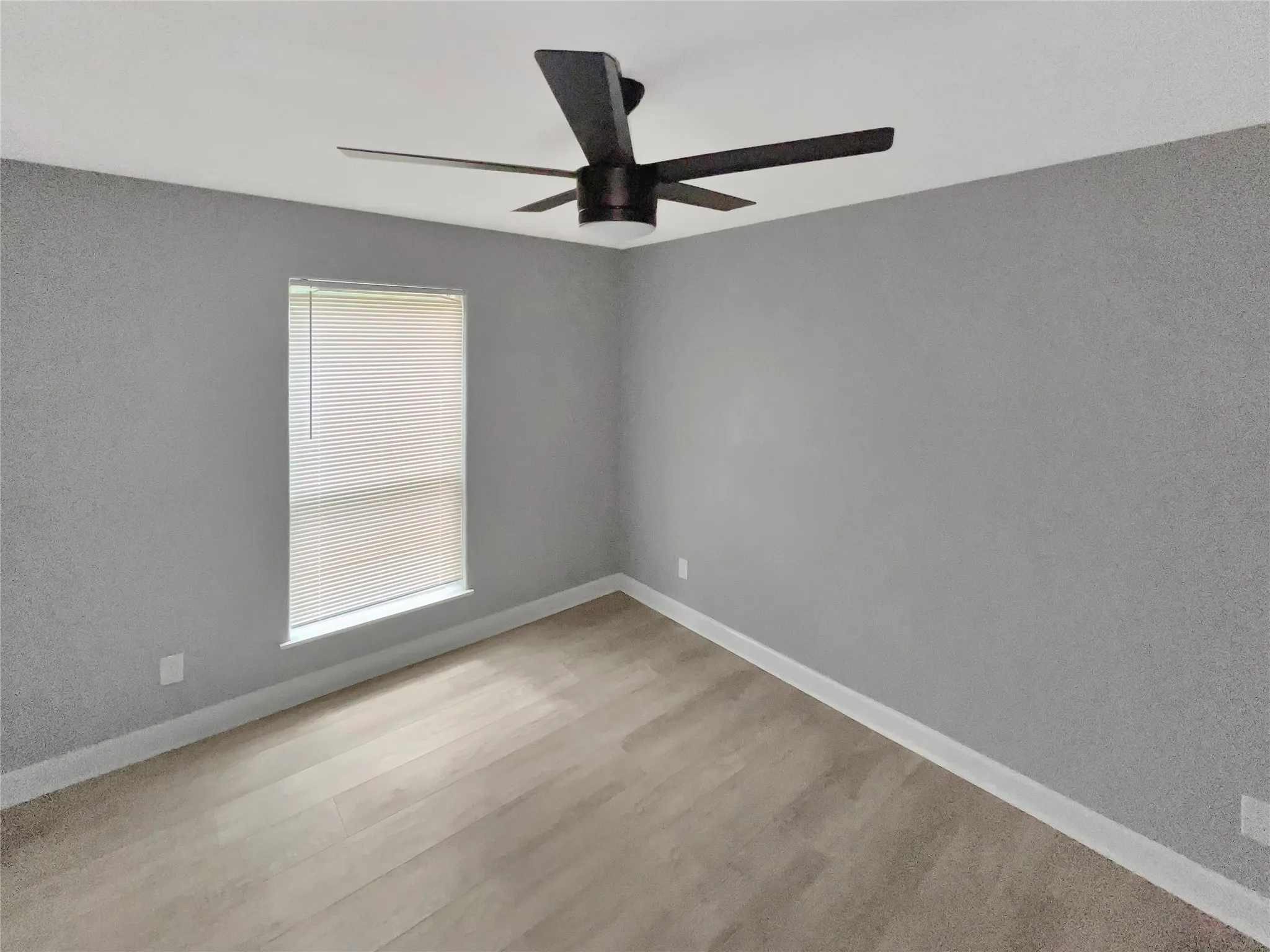 Room featuring wood-finish flooring, a window with blinds, a ceiling fan with dark blades, and light gray walls with white baseboards