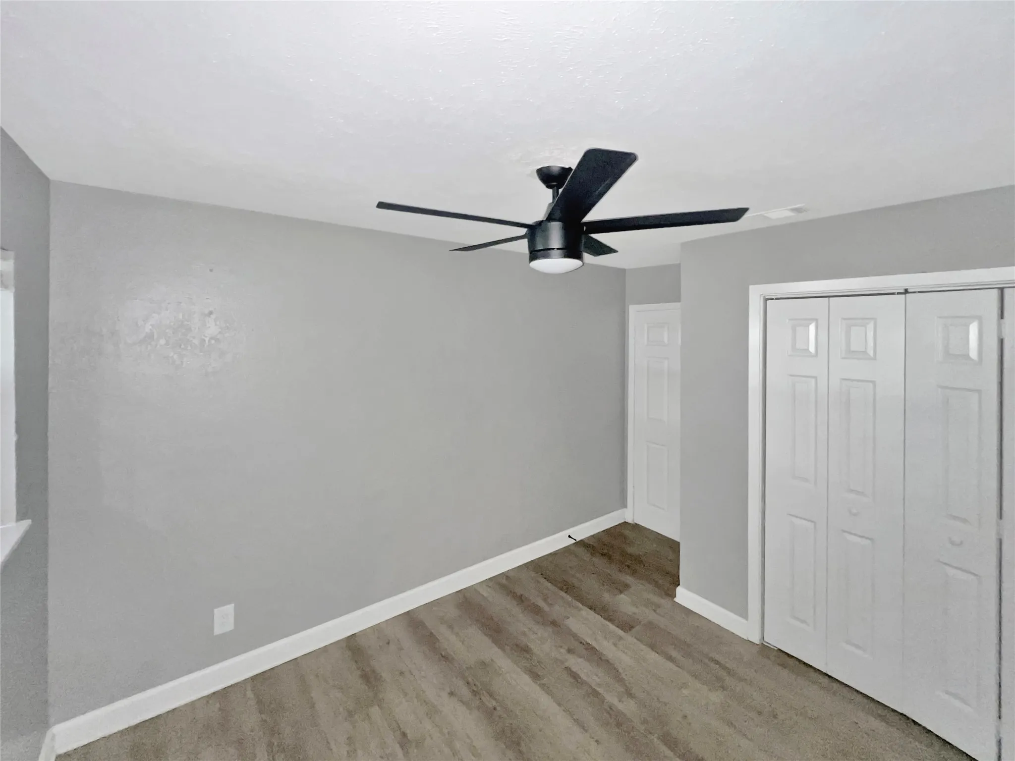 Room featuring wood-finish flooring, light grey painted walls, white baseboards, and a contemporary black ceiling fan with integrated lighting