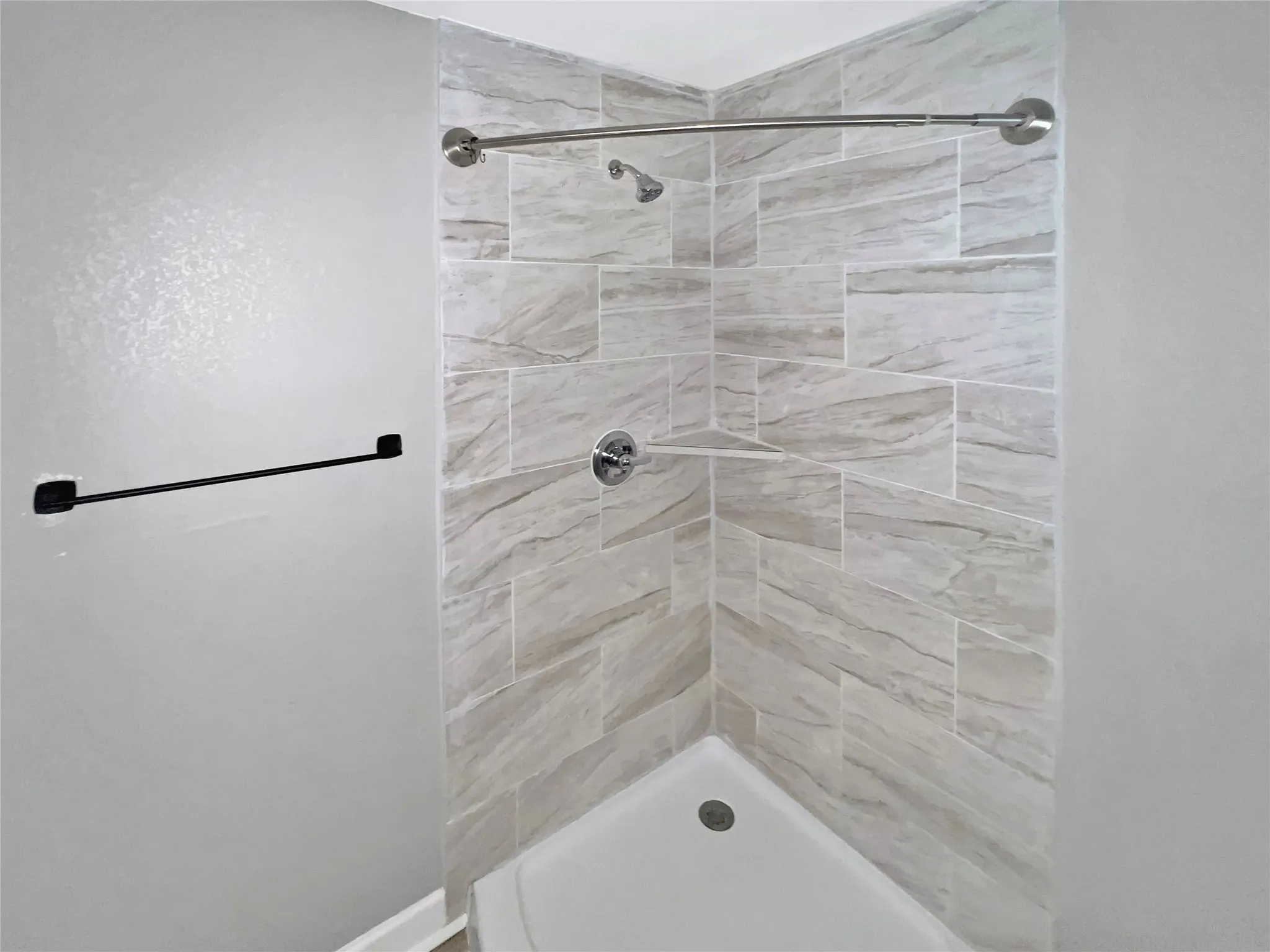 Corner shower featuring light-toned tilework with a varied pattern, a built-in shower niche, and a contrasting matte black towel bar