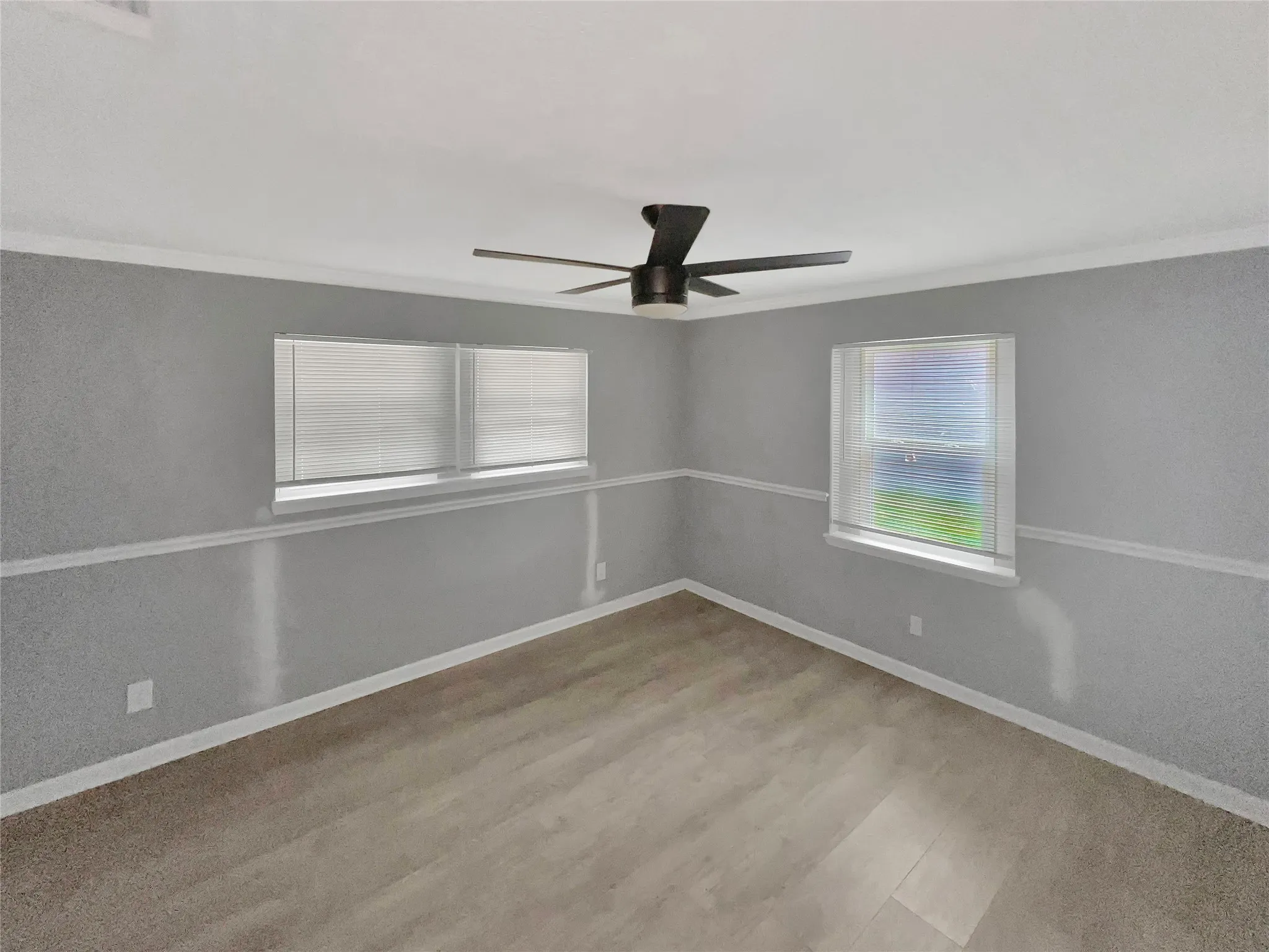 Spacious room featuring wood-finish flooring, two windows with blinds, a modern ceiling fan, and grey walls with white trim