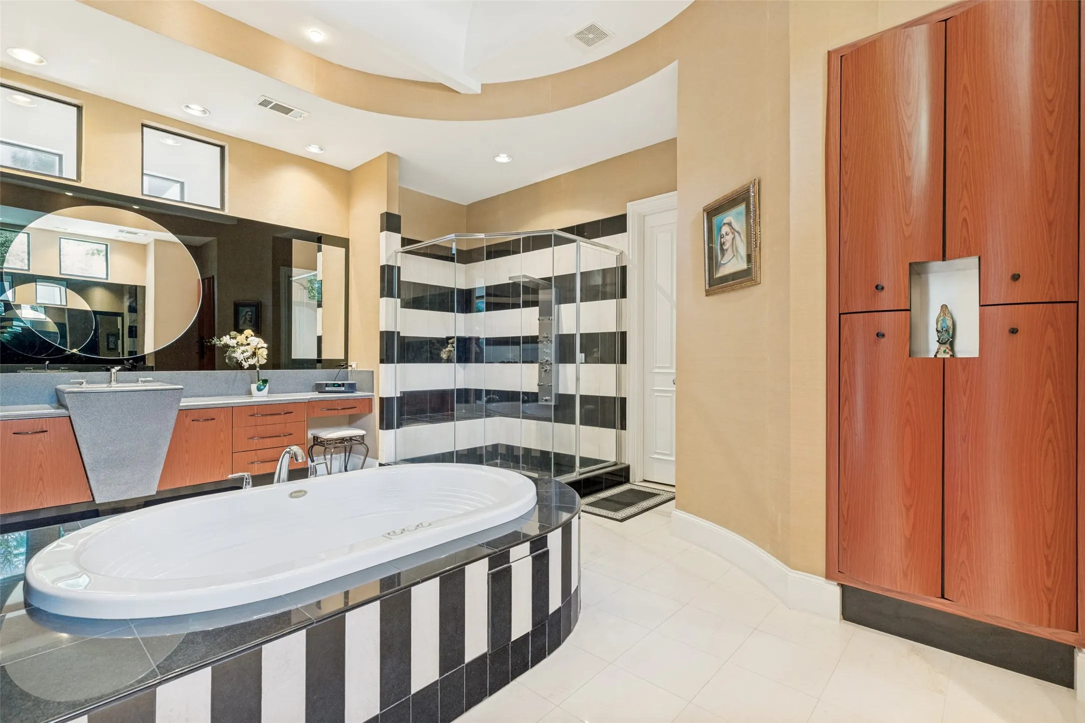 Spacious bathroom featuring a white soaking tub with a black and white striped base, a glass-enclosed shower with matching black and white striped tiling, a dual vanity with a unique gray basin and wood-finish cabinetry, and built-in wood-finish storage