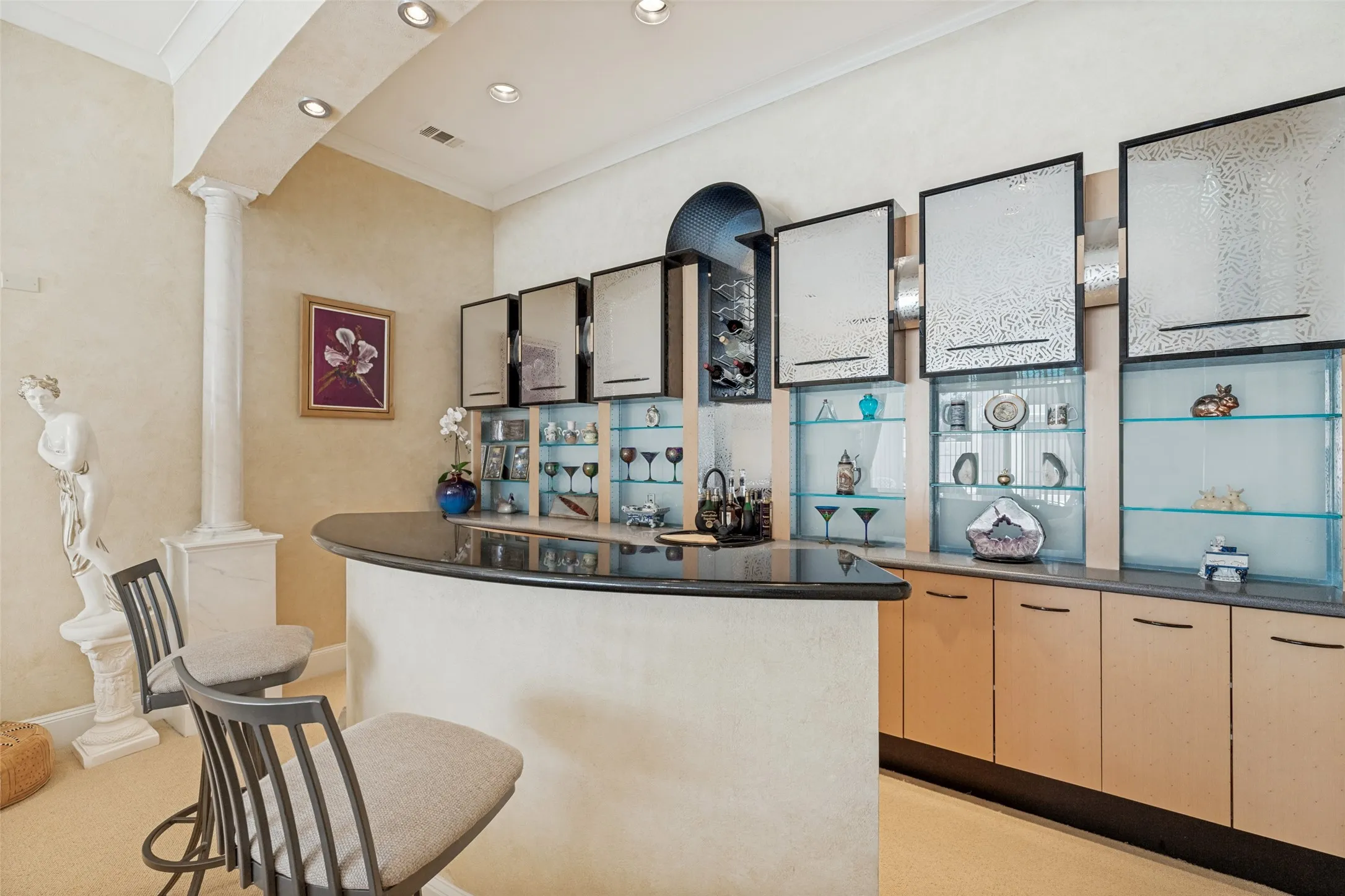Curved bar with a dark countertop and integrated shelving, featuring glass-front display cabinets, light wood-finish lower cabinetry, and recessed lighting