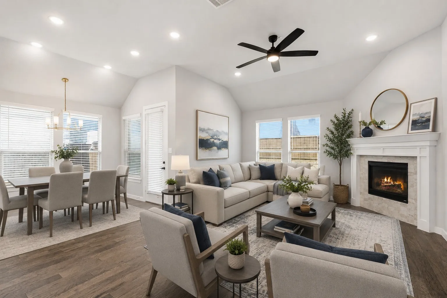 Open-concept living area featuring vaulted ceilings, recessed lighting, wood-finish flooring, a corner fireplace with a white mantel, and multiple windows with blinds