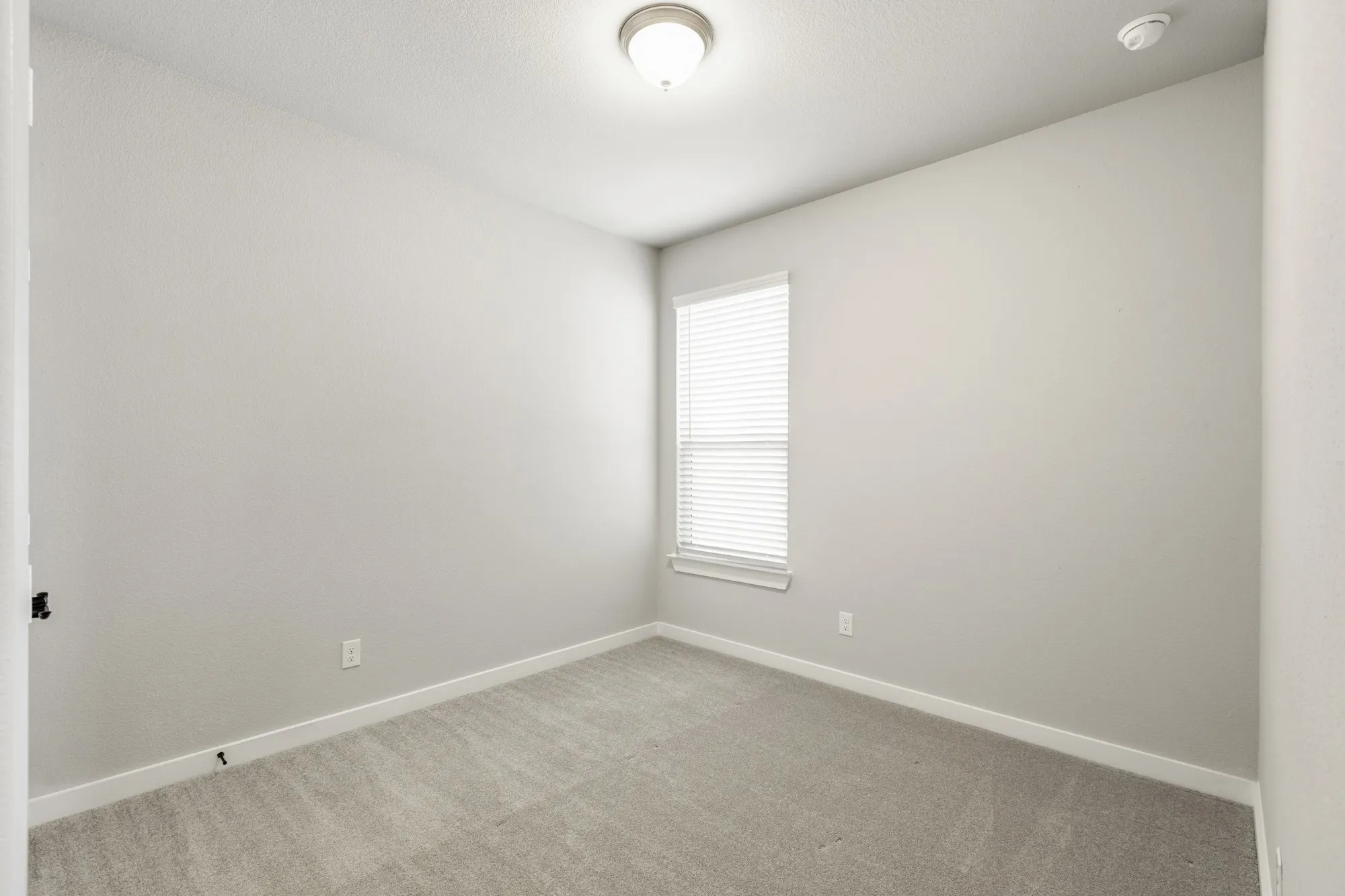 Carpeted room with a single window featuring blinds, a ceiling-mounted light fixture, and light-toned walls