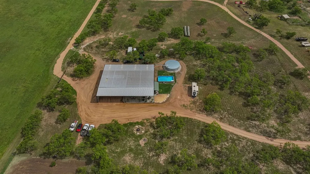 Aerial view of the ranch homestead showing the barndominium, pool, 40,000 gallon rainwater tank, firepit and RV parking. Shady mesquite trees surround it and hay fields sit just beyond.