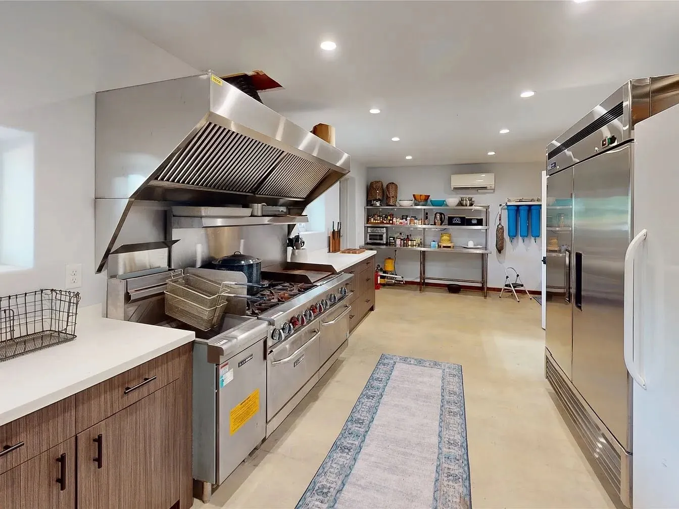 Commercial kitchen with a six burner range, griddle, double ovens, fryer and a stainless vent hood. A two door reach in refrigerator and pantry shelving round it out, ready for hunting lodge meals, retreat catering or event venue use.