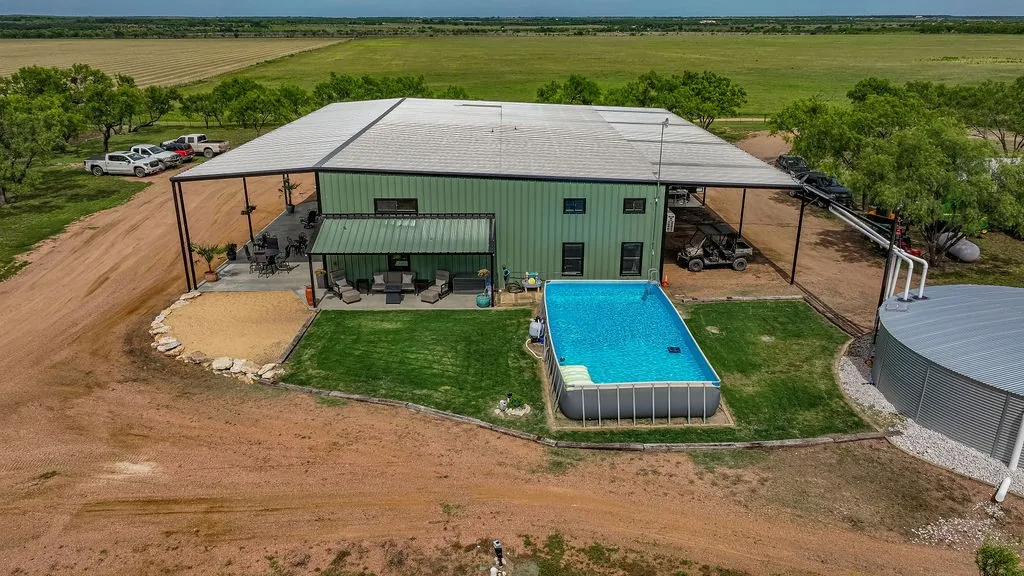 Aerial of the barndominium backyard: a fenced lawn, above ground pool, covered patio and attached carport parking. Productive hay fields and native mesquite stretch beyond.