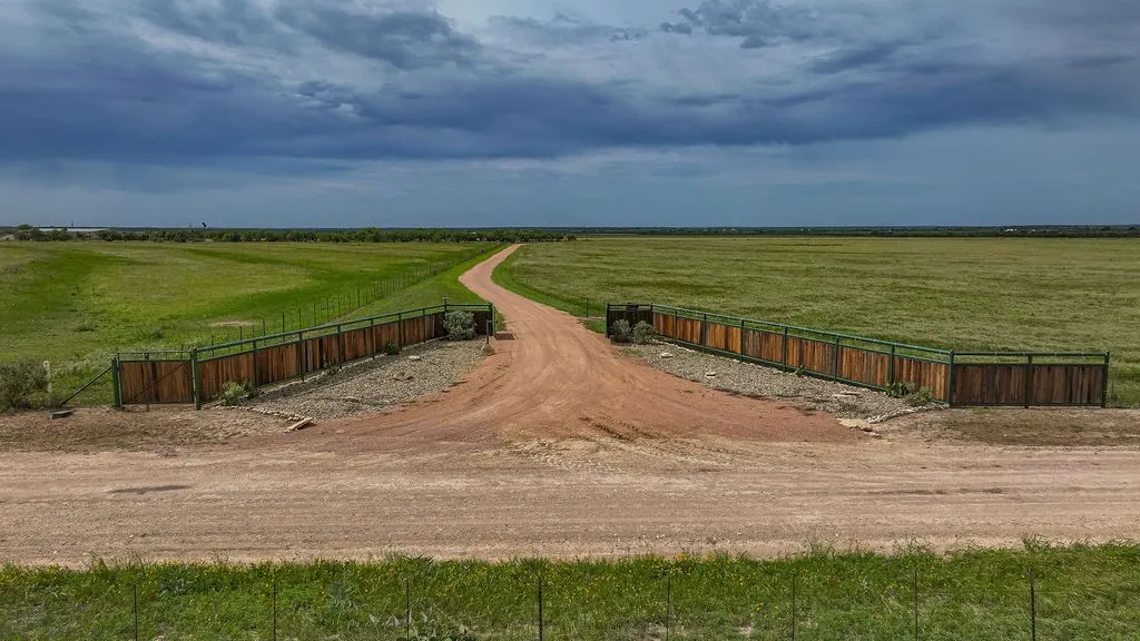 Private ranch entrance on CR 419 with custom steel and cedar entry fencing. A wide caliche drive leads through open coastal Bermuda pasture, ideal for a cattle ranch, hay operation or horse property.