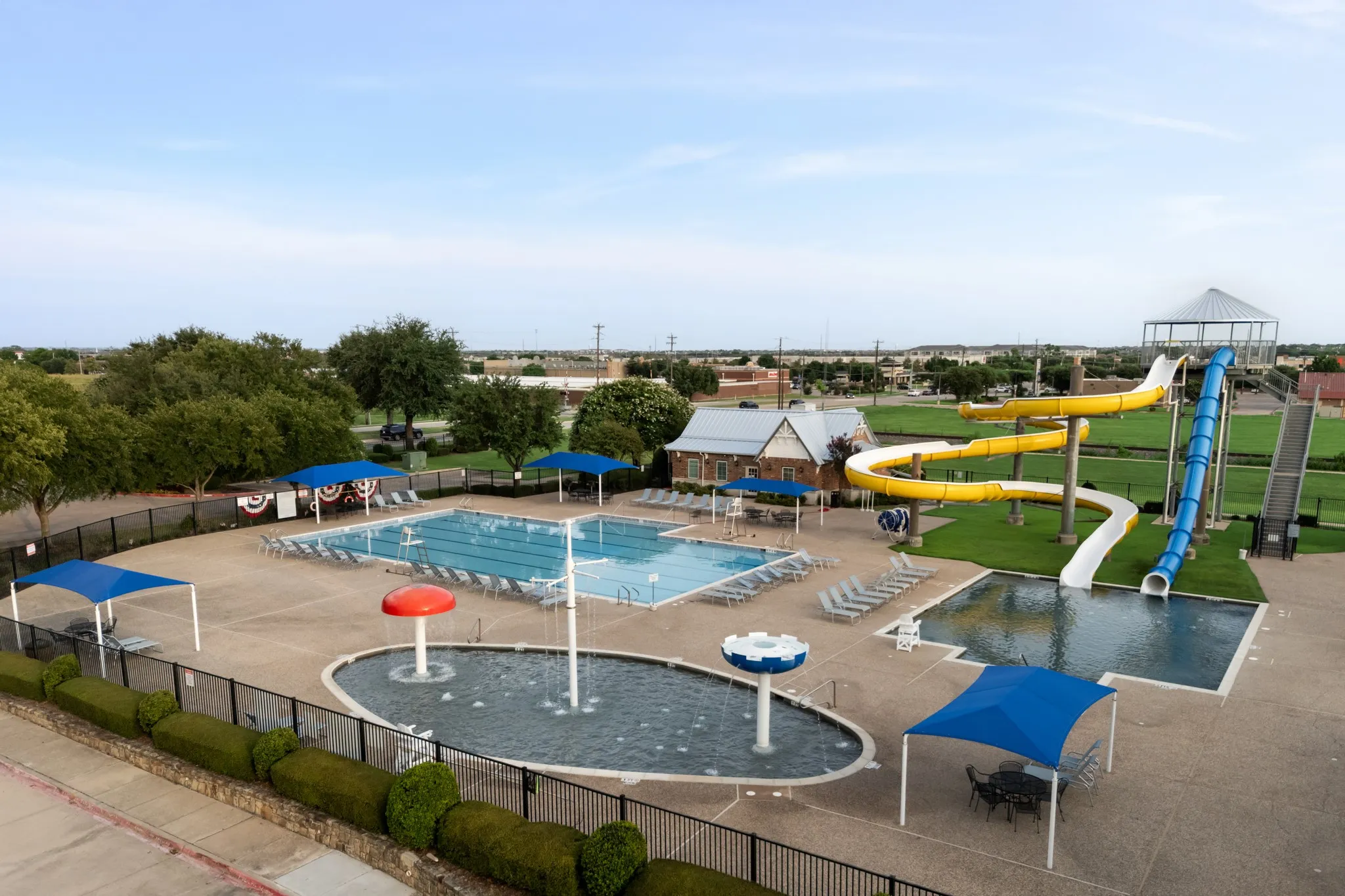Community pool area featuring a large swimming pool, a separate splash pad with water features, multiple lounge chairs, and a recreational water slide structure
