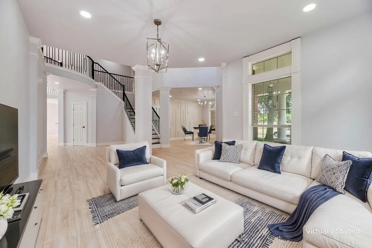 Expansive living area featuring light wood-finish flooring, a grand staircase with wrought iron balusters, a decorative chandelier, recessed lighting, and a large multi-pane window