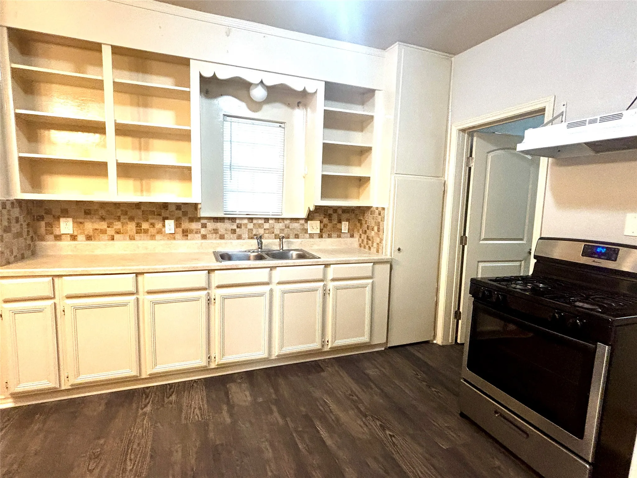 Kitchen featuring a stainless steel gas range, wood-finish flooring, mosaic tile backsplash, double basin sink, and ample white cabinetry with open shelving