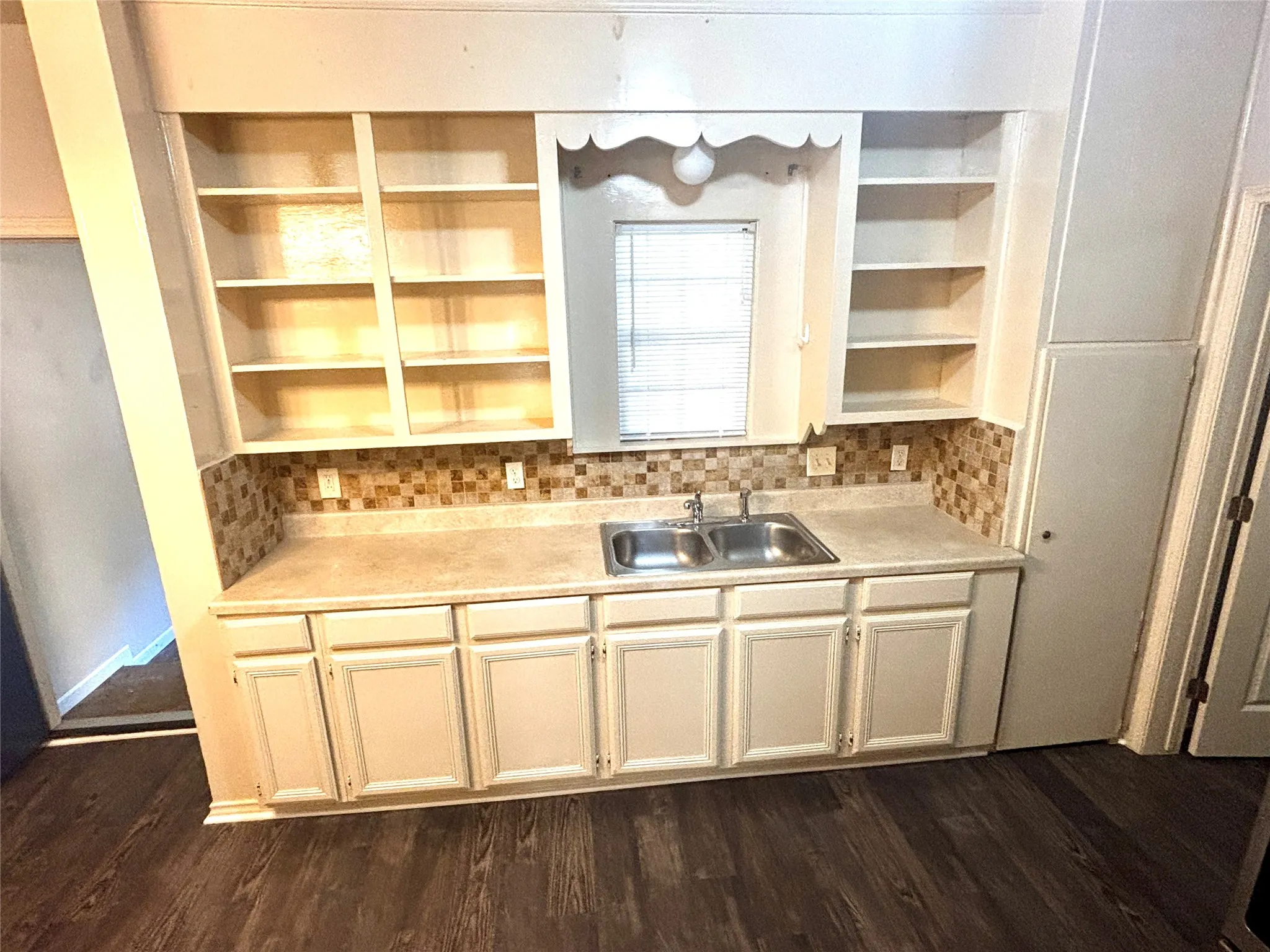 Kitchenette featuring wood-finish flooring, light-toned base cabinetry, open shelving, a double basin stainless steel sink, and a tiled backsplash