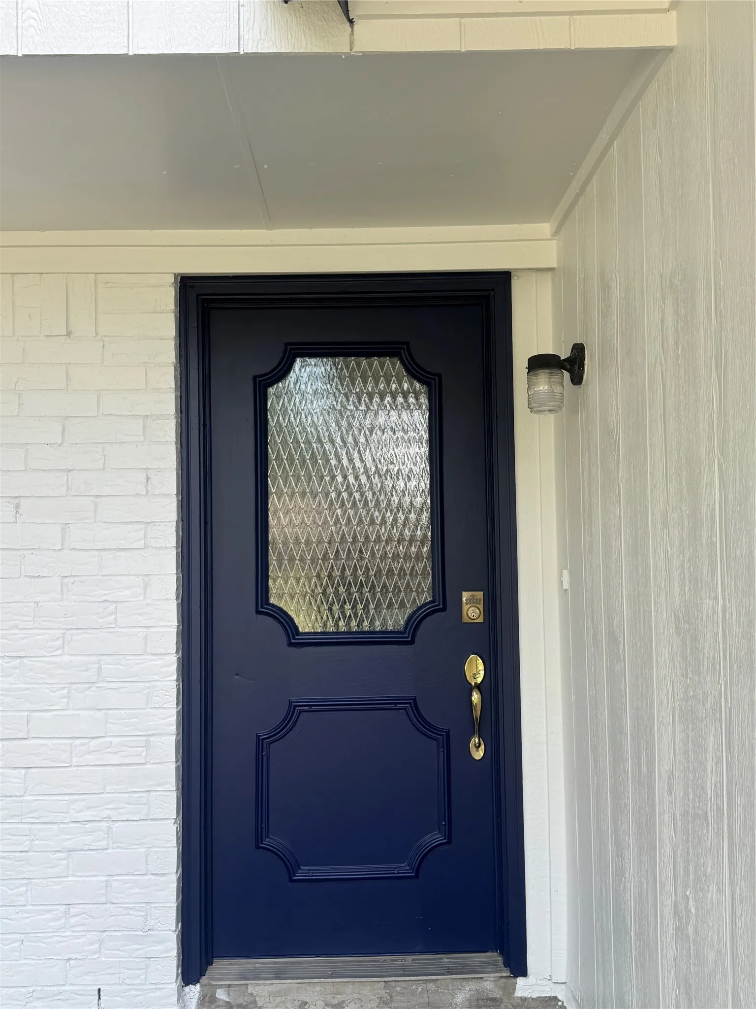Navy blue entry door with decorative glass insert, bright brass hardware, painted brick siding, and vertical wood paneling