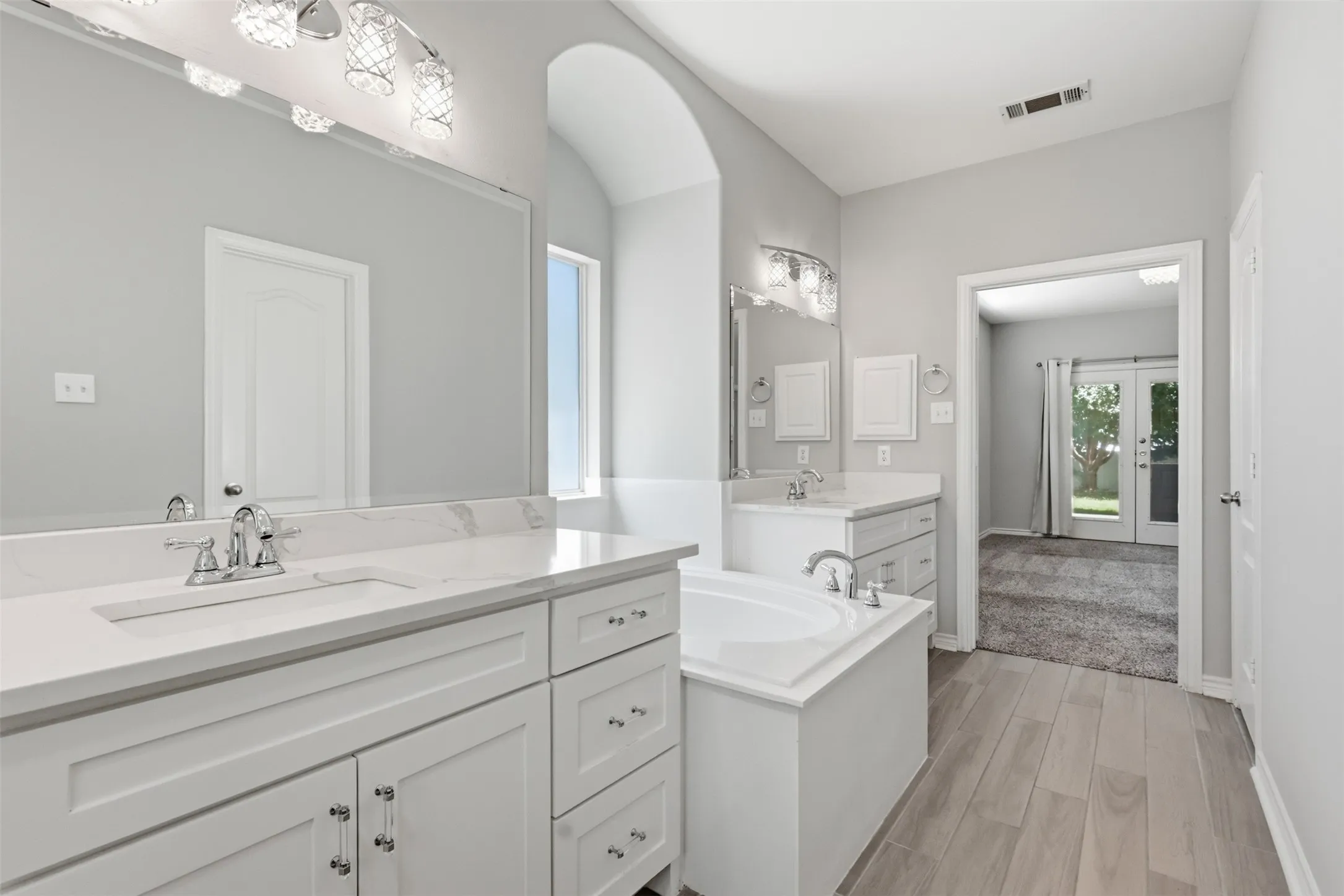 Dual vanity bathroom featuring light-toned wood-finish flooring, white cabinetry with chrome hardware, an undermount tub, and an arched entryway