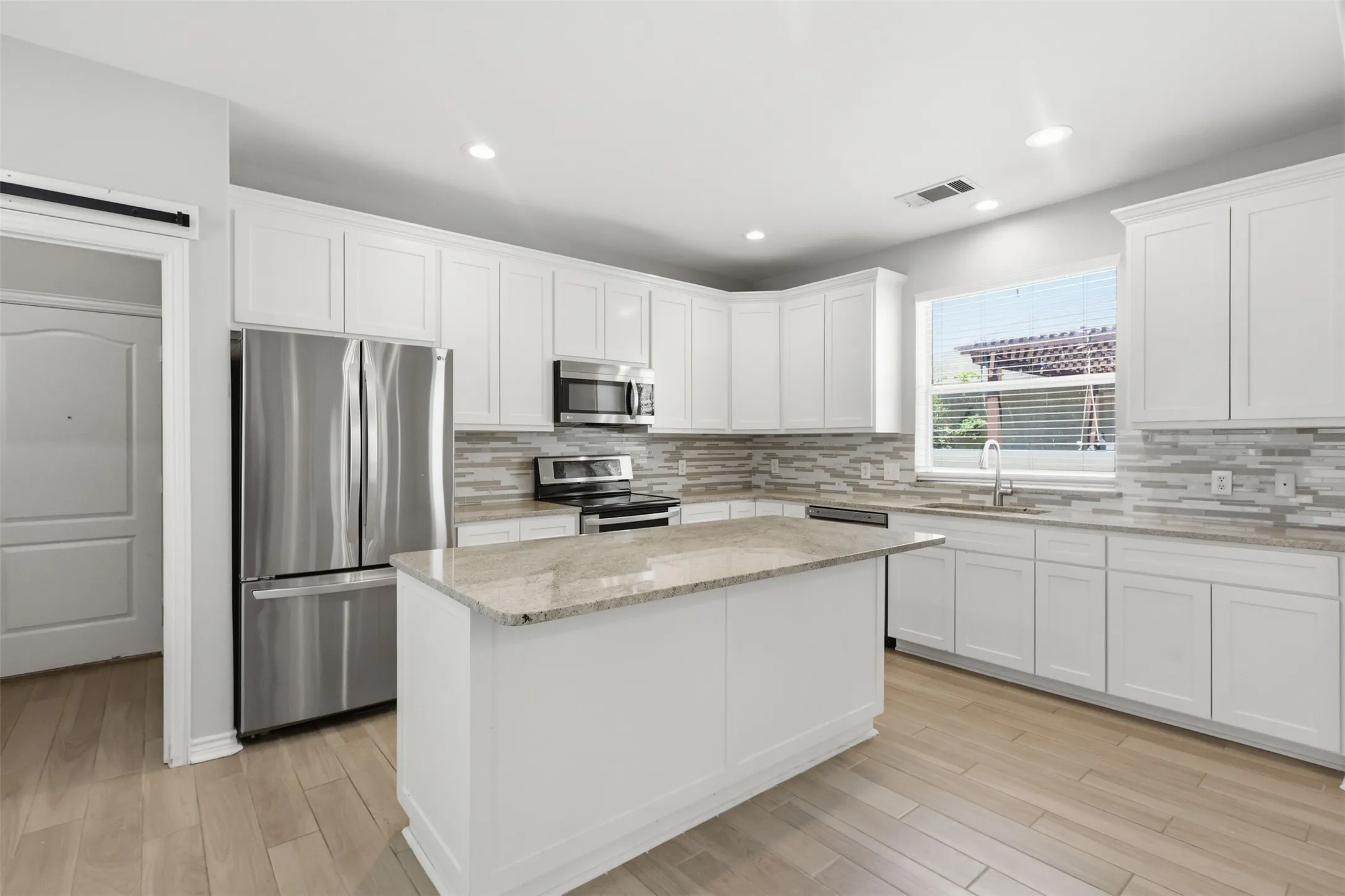 Bright kitchen featuring white cabinetry, light wood-finish flooring, a central island with a stone countertop, a tile backsplash, and stainless steel appliances