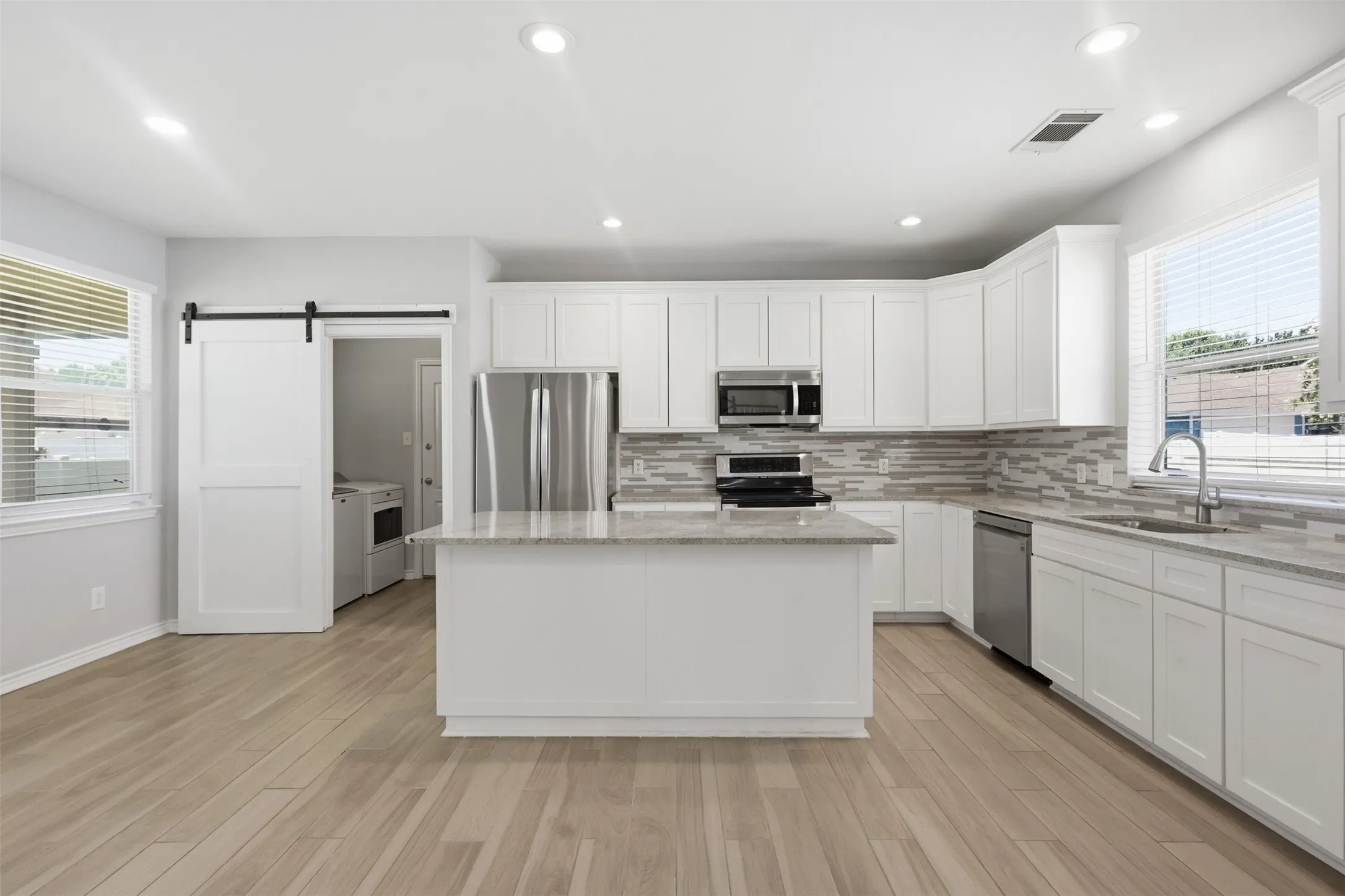 Open-concept kitchen featuring an island with a light-toned countertop, white cabinetry, stainless steel appliances, a multi-tone tile backsplash, and wood-finish flooring