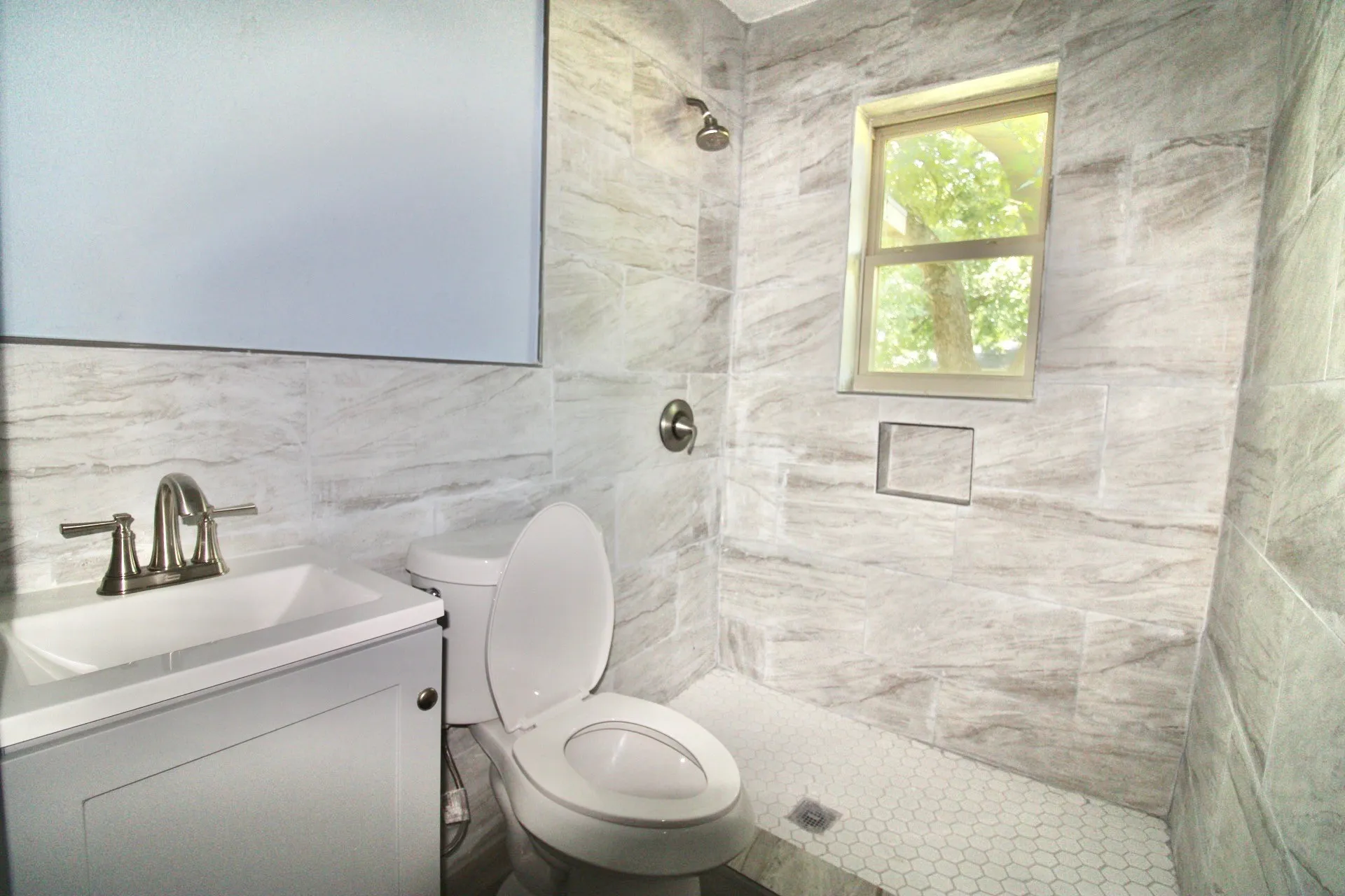 Contemporary bathroom featuring large format stone-look tile walls, white hexagonal mosaic shower floor, and a built-in shower niche