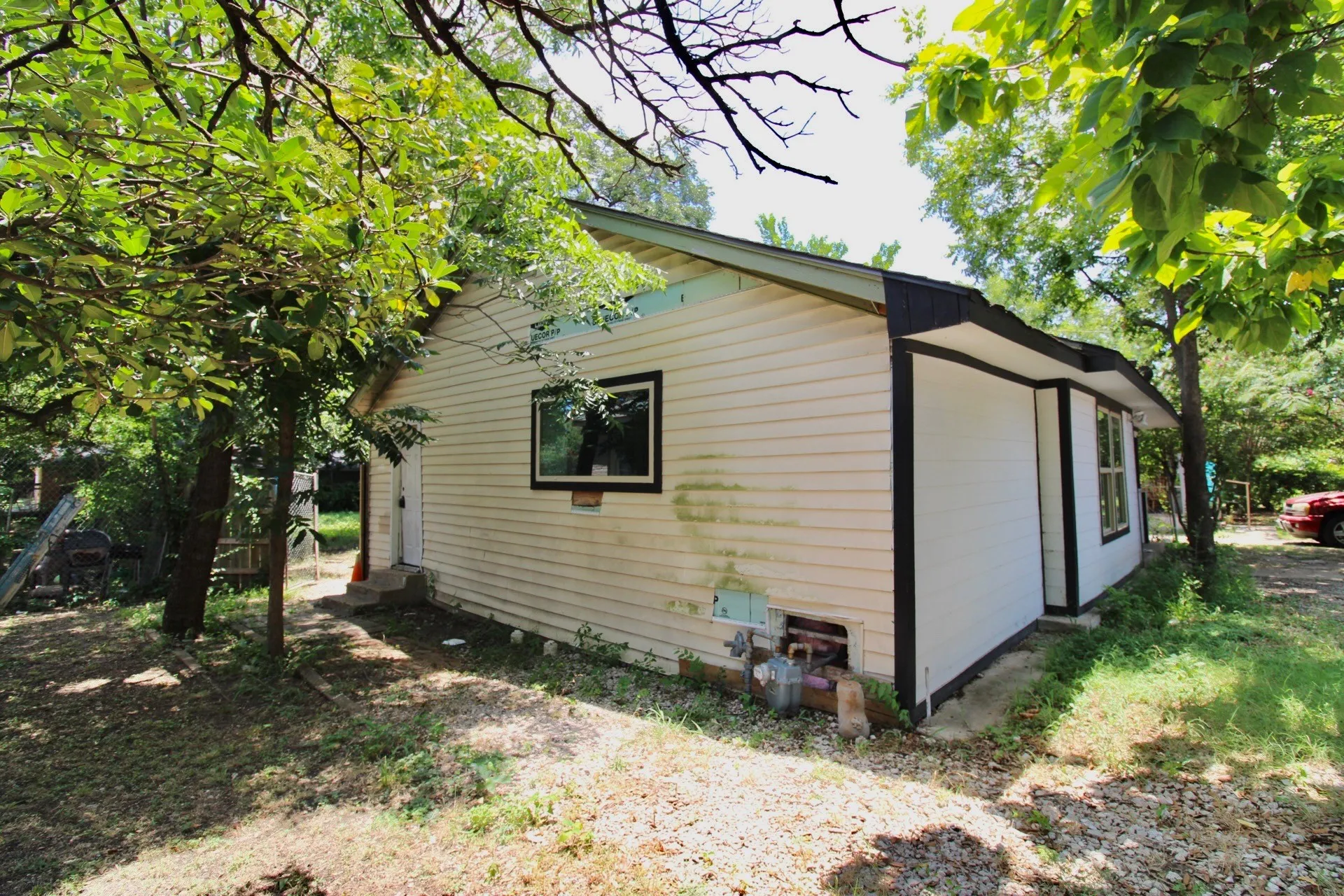 Exterior siding featuring a window with black trim, a gravel and grass ground covering, and surrounding mature trees