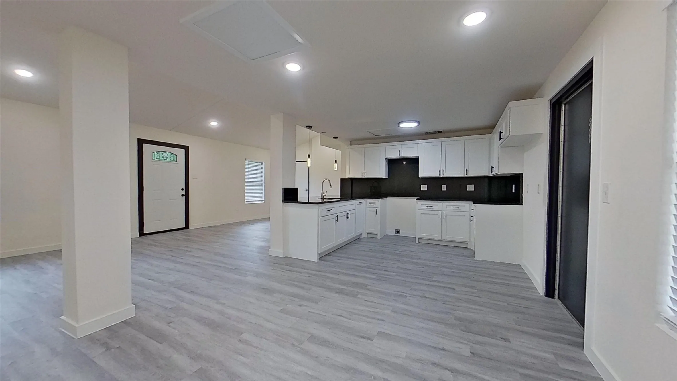 Open-concept living area featuring light wood-finish flooring, recessed lighting, and a contemporary kitchen with white shaker cabinetry, dark countertops, and a full-height dark backsplash