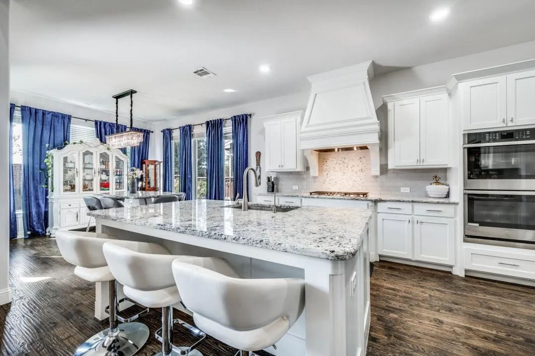 Spacious kitchen featuring a large island with a light-toned countertop, white cabinetry, stainless steel double ovens, and a decorative range hood