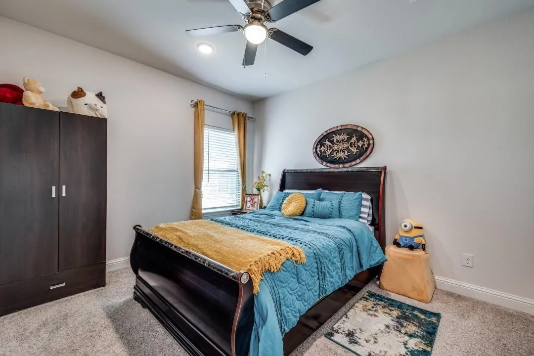 Carpeted bedroom featuring a window with blinds and drapes, a dark wood armoire, a built-in light fixture, and a ceiling fan with integrated lighting