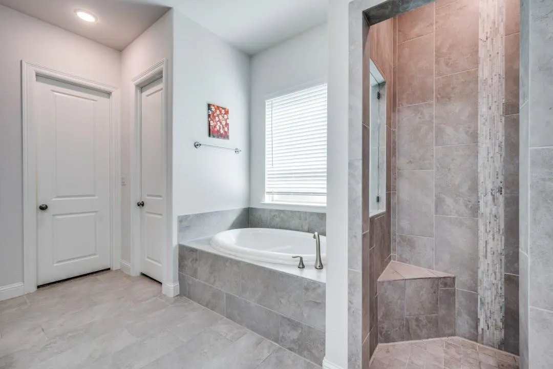 Bathroom featuring large format tile flooring, a soaking tub with tile surround, a window with blinds, and a walk-in shower with a tiled bench and decorative tile accent
