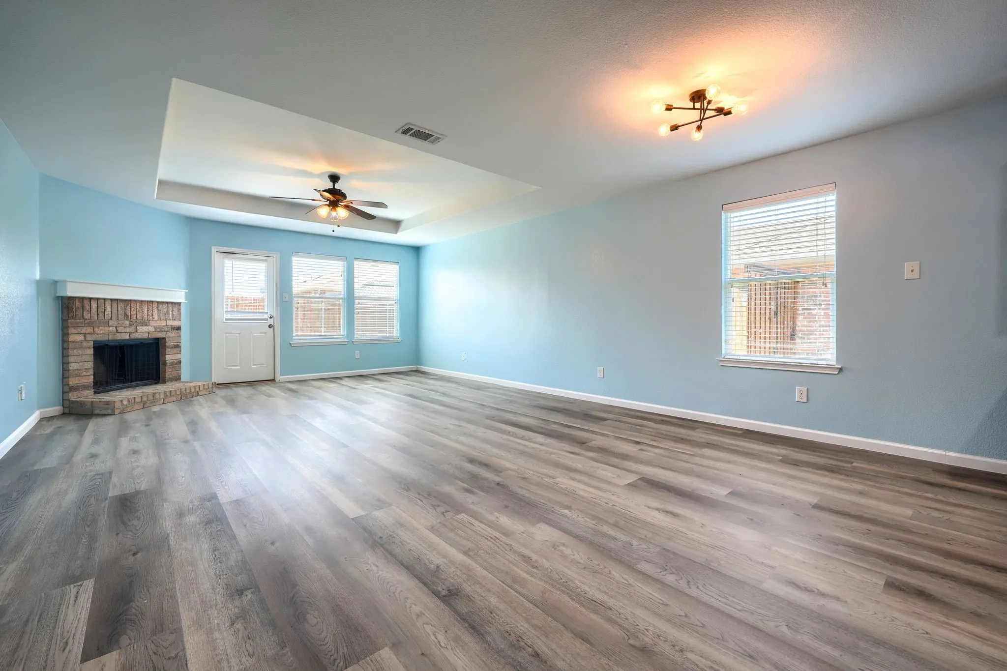 Spacious room featuring wood-finish flooring, a corner brick fireplace with a white mantel, and light blue walls