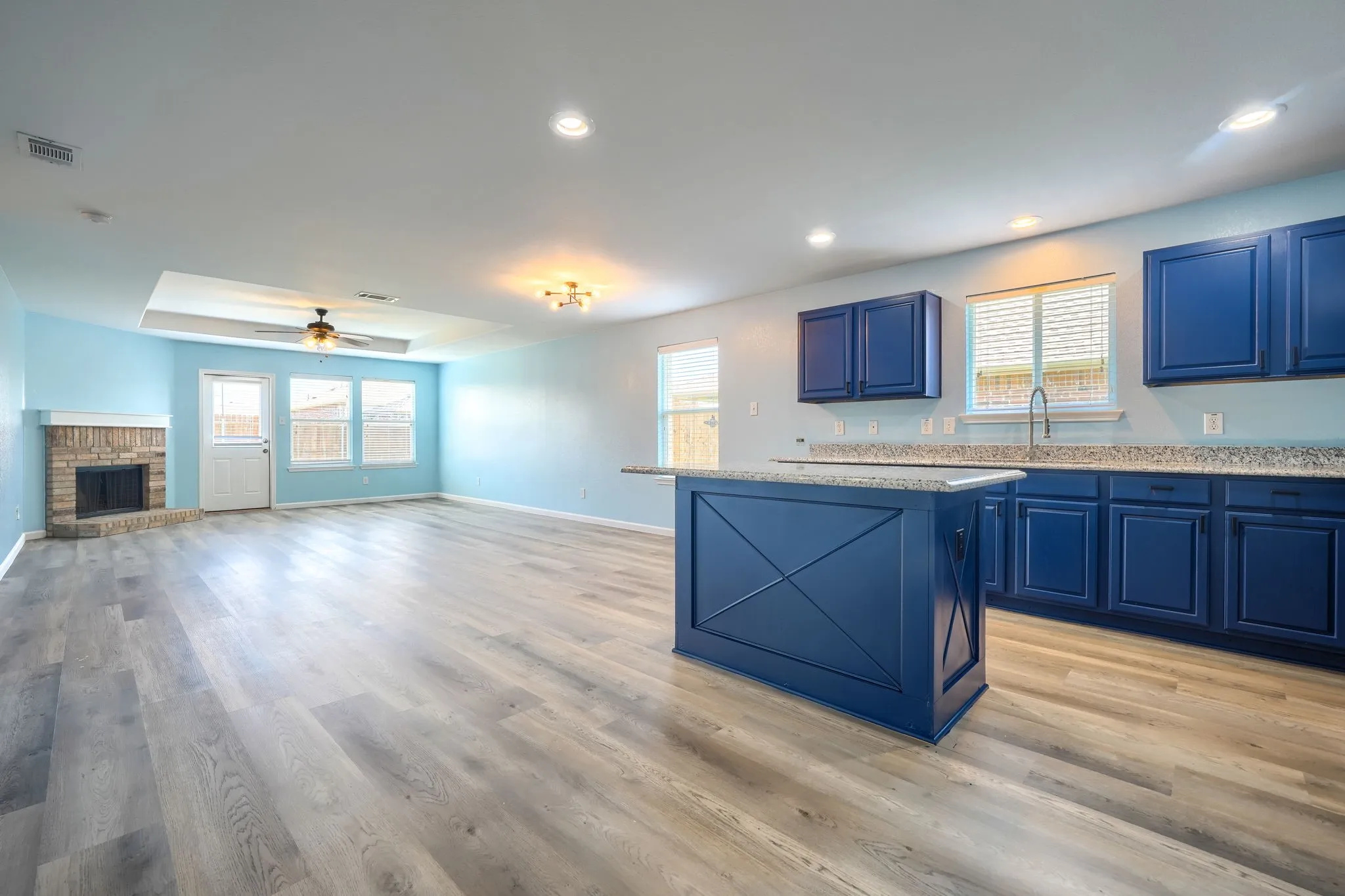 Open-concept living space featuring wood-finish flooring, a brick fireplace, a tray ceiling with a ceiling fan, and a kitchen island with granite-style countertops and navy cabinetry