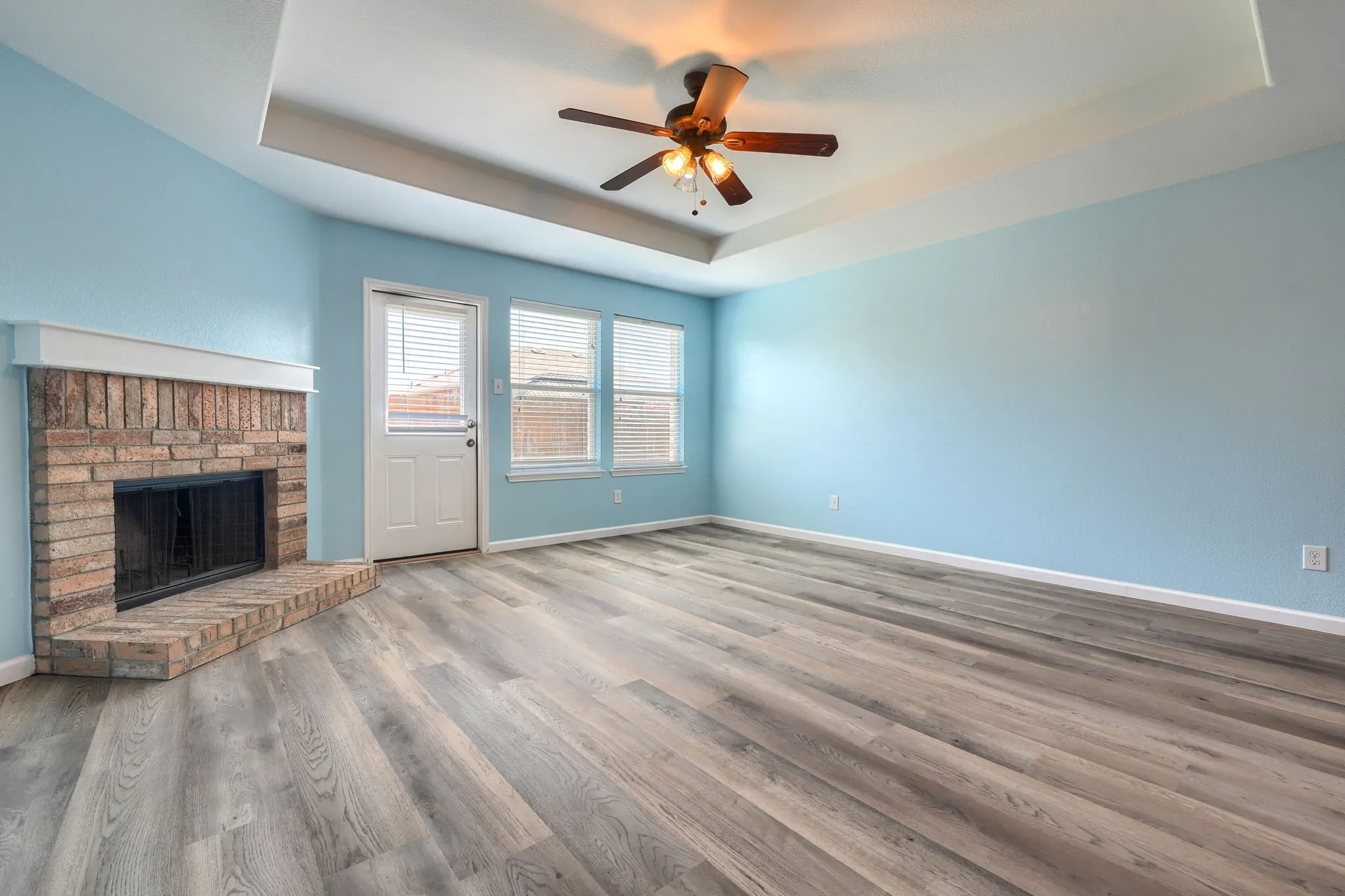 Spacious living area featuring a corner brick fireplace with hearth, wood-finish flooring, a ceiling fan, and a tray ceiling