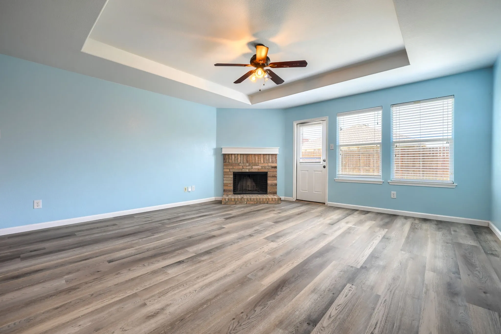 Spacious room featuring wood-finish flooring, a tray ceiling with a ceiling fan, and a brick-surround fireplace